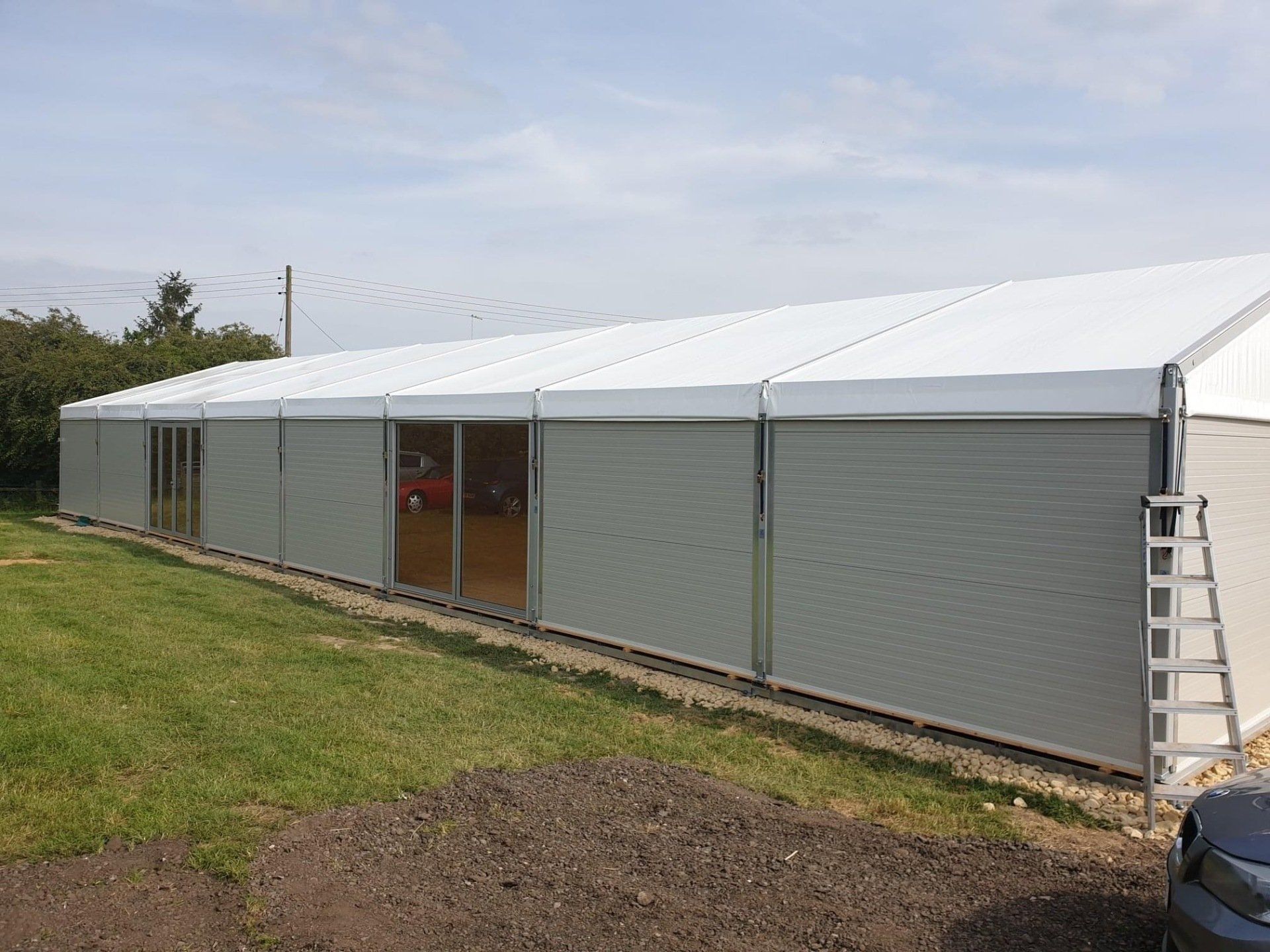 A long, gray temporary structure with a white roof on a grassy area, a ladder on the right.