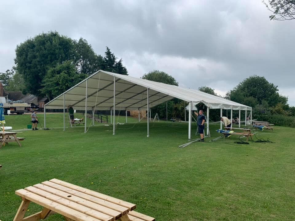 People assembling a white tent on a green grassy area with picnic tables. Cloudy sky in the background.