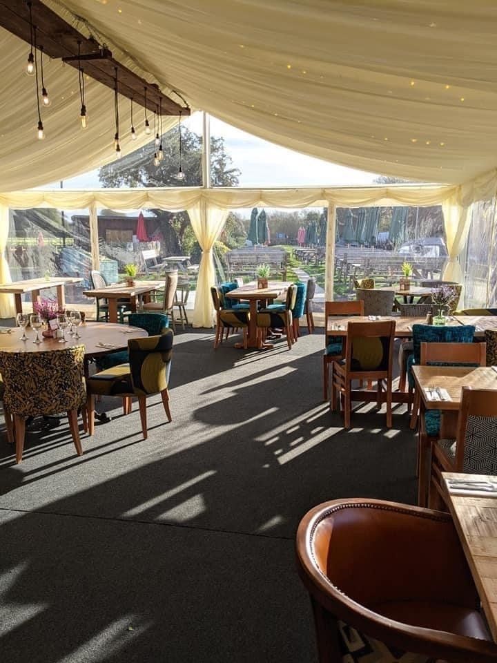 Interior of a tent restaurant with tables, chairs, and large windows. Sunlight streams in, illuminating the space.