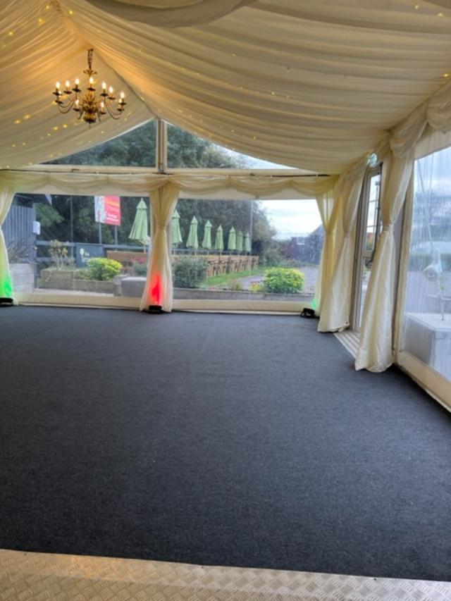 Inside a tent with black carpet, clear walls, and a chandelier, overlooking a garden.