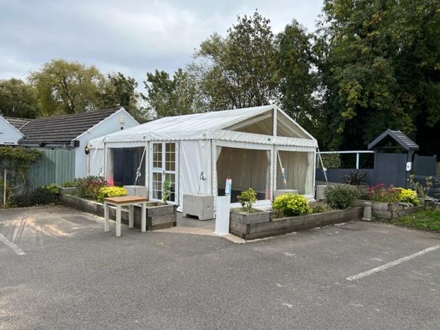 White tent with clear side panels, next to a building and landscaping, in a parking area.