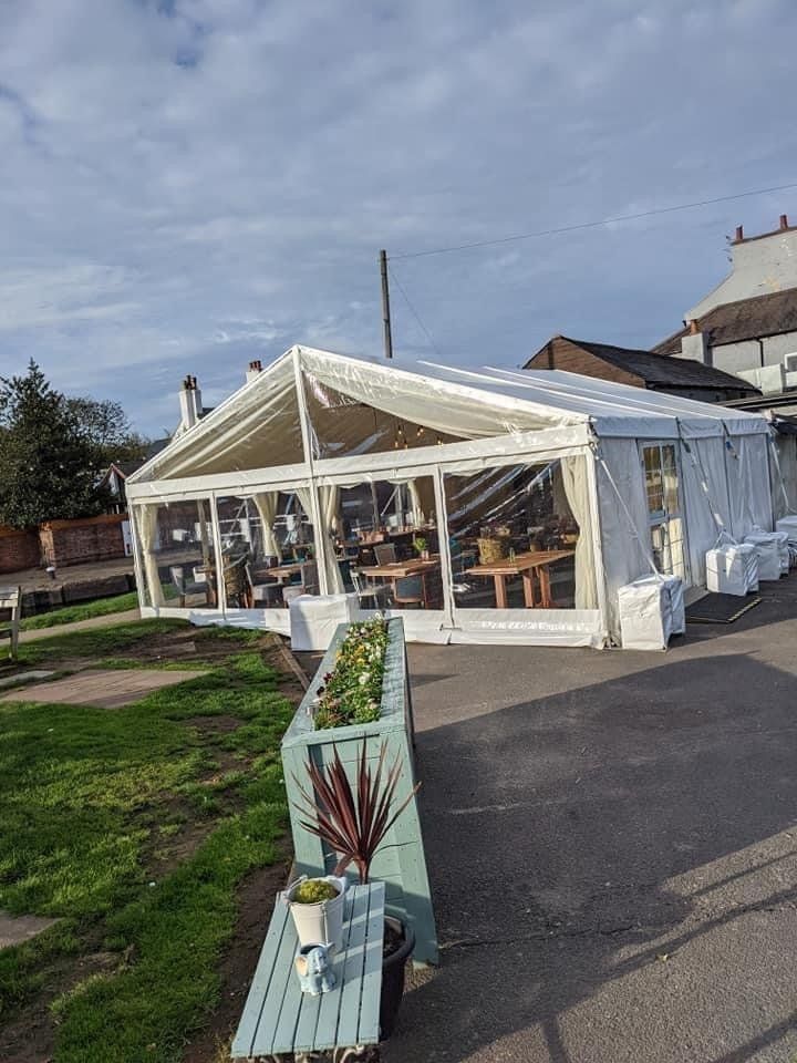 A white tent with clear windows, set up outside a building. Tables and chairs visible inside.