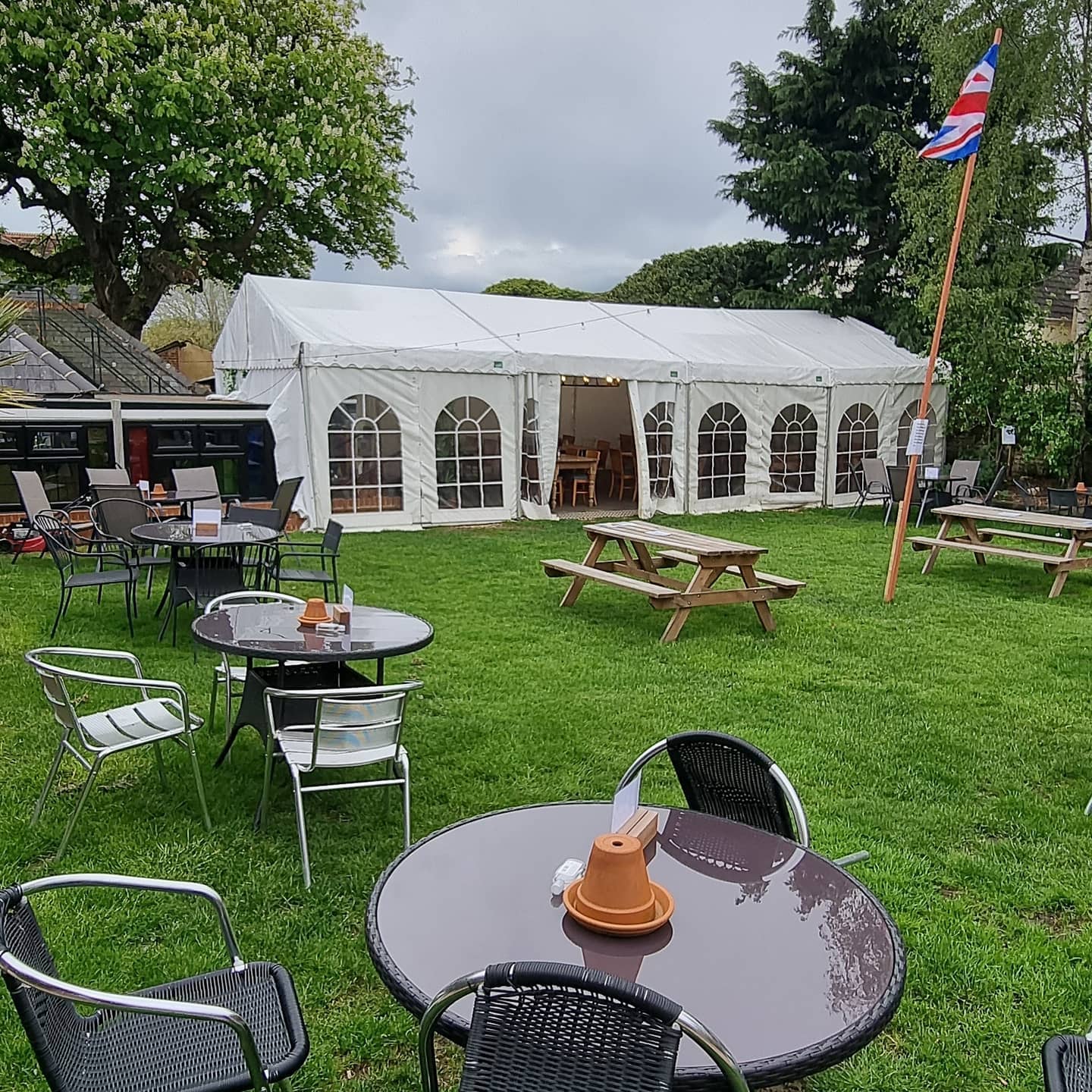 Outdoor dining area with white tent, tables, chairs, and picnic benches on green grass.