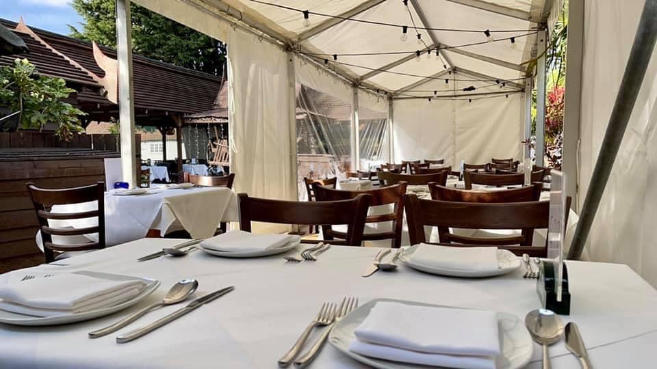 Restaurant tent with white-covered tables, wooden chairs, and place settings. Exterior view with other structures visible.