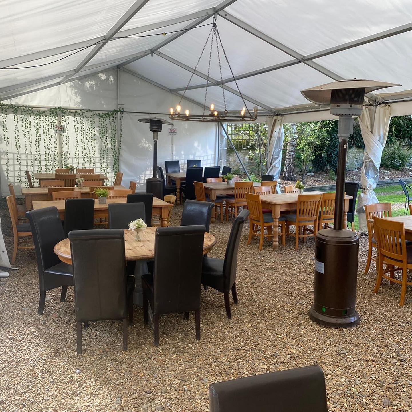 Tent-covered outdoor dining area with tables, chairs, two heaters, and a chandelier.