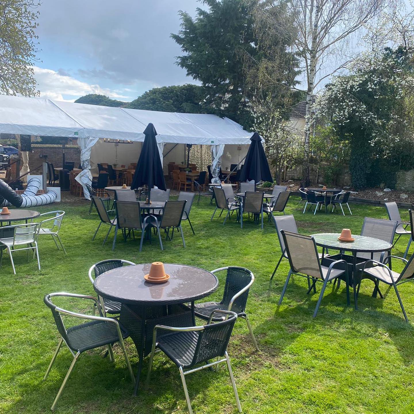 Outdoor seating area with tables and chairs on a grassy lawn. Tents and umbrellas provide shade.