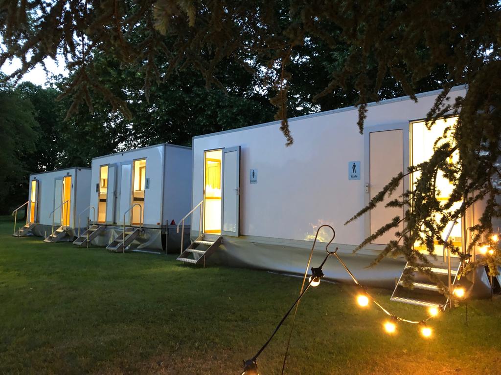 Row of illuminated white portable restroom units on grass, strung with lights, in a wooded outdoor setting.