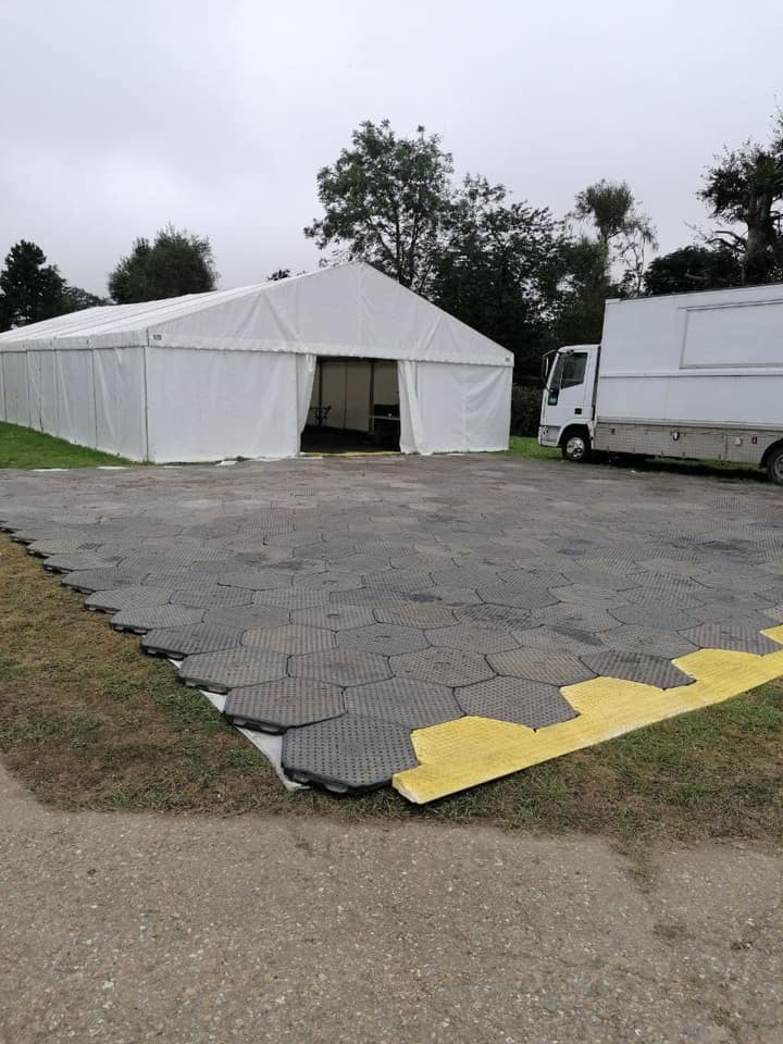A large white tent with open entrance, parked next to a truck. Dark interlocking tiles cover the ground. Cloudy sky.