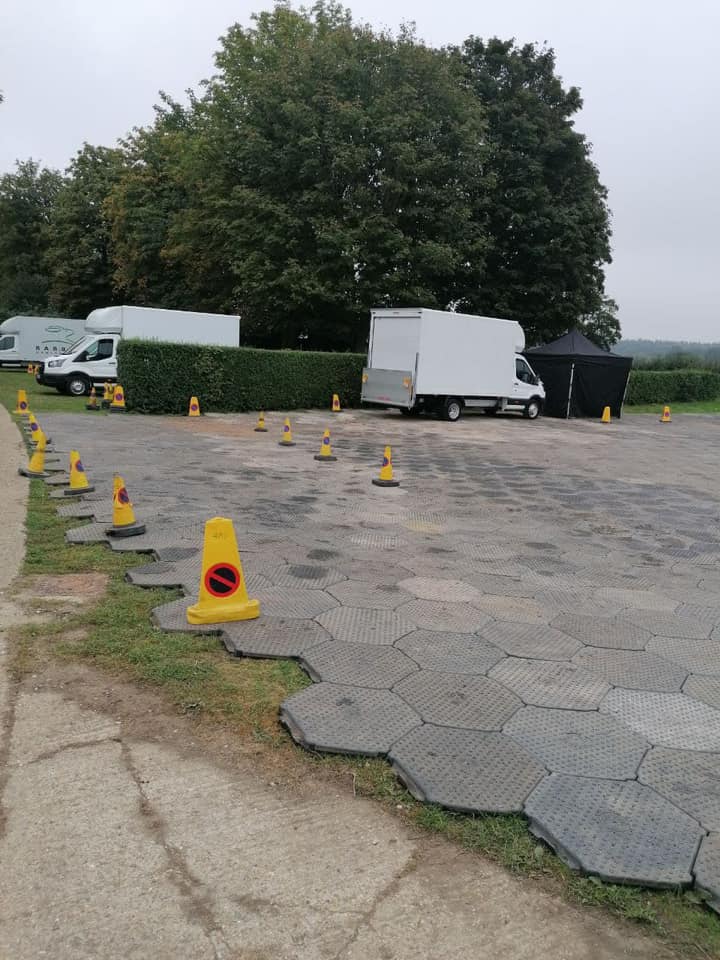 Parking area with trucks and traffic cones, next to a hedge and trees, on a cloudy day.