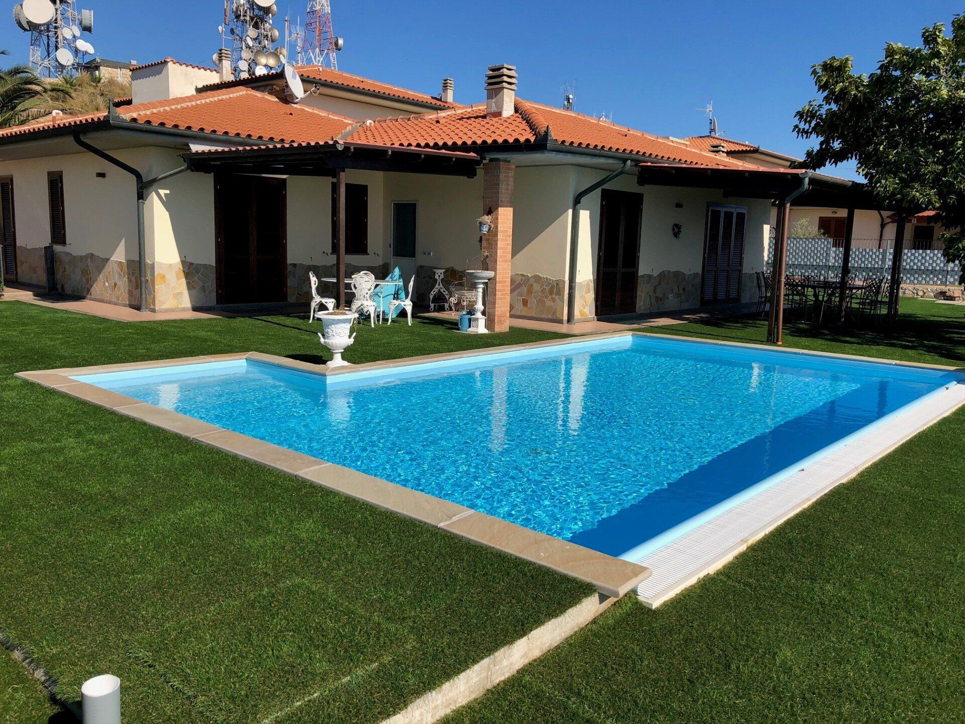 Un cortile soleggiato con una piscina rettangolare, un prato verde lussureggiante e una casa color crema con tetto in terracotta.