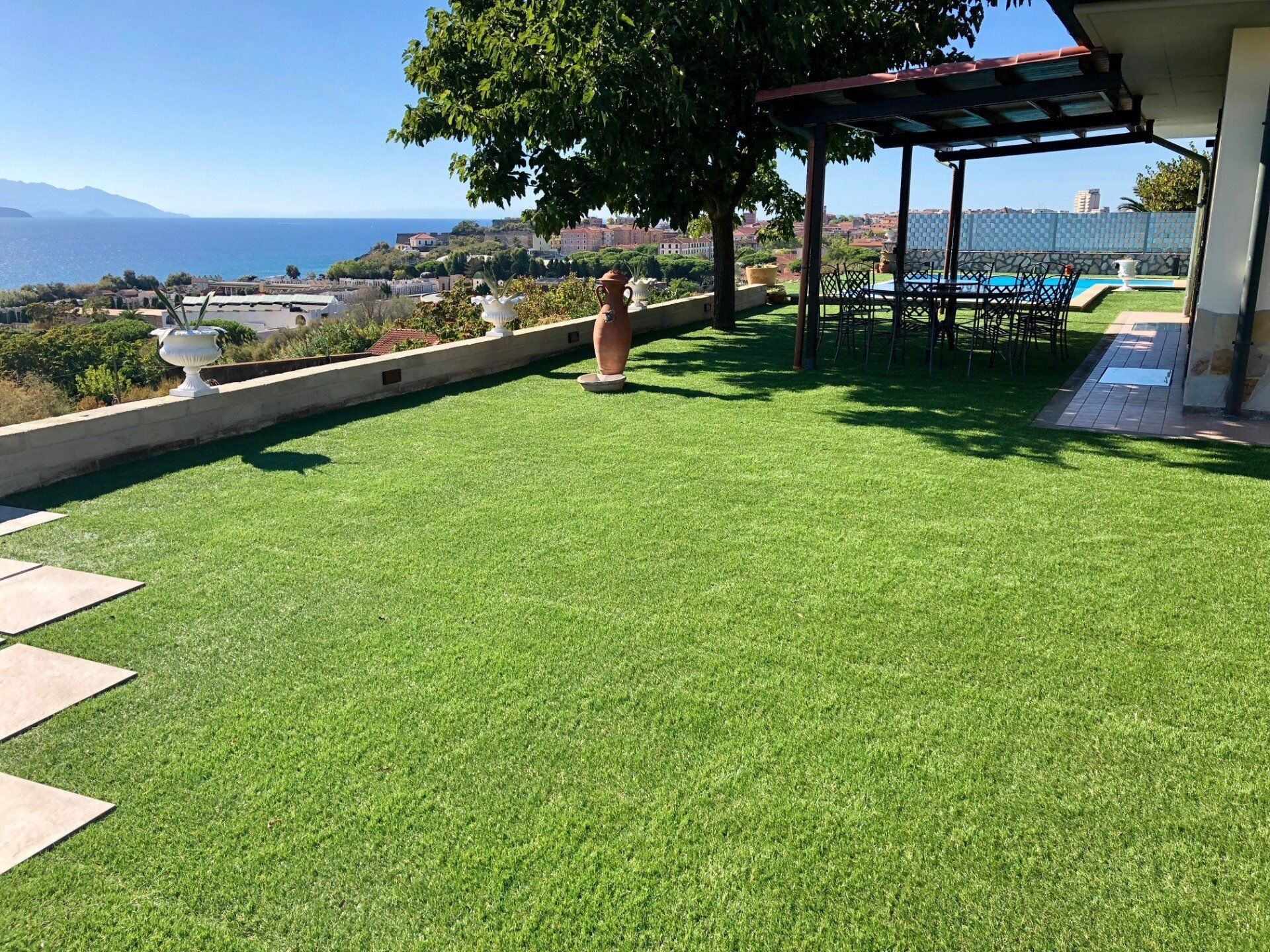 Un patio erboso sul retro della casa, con un pergolato, una pianta in vaso e una vista panoramica sul mare blu e sulla costa.