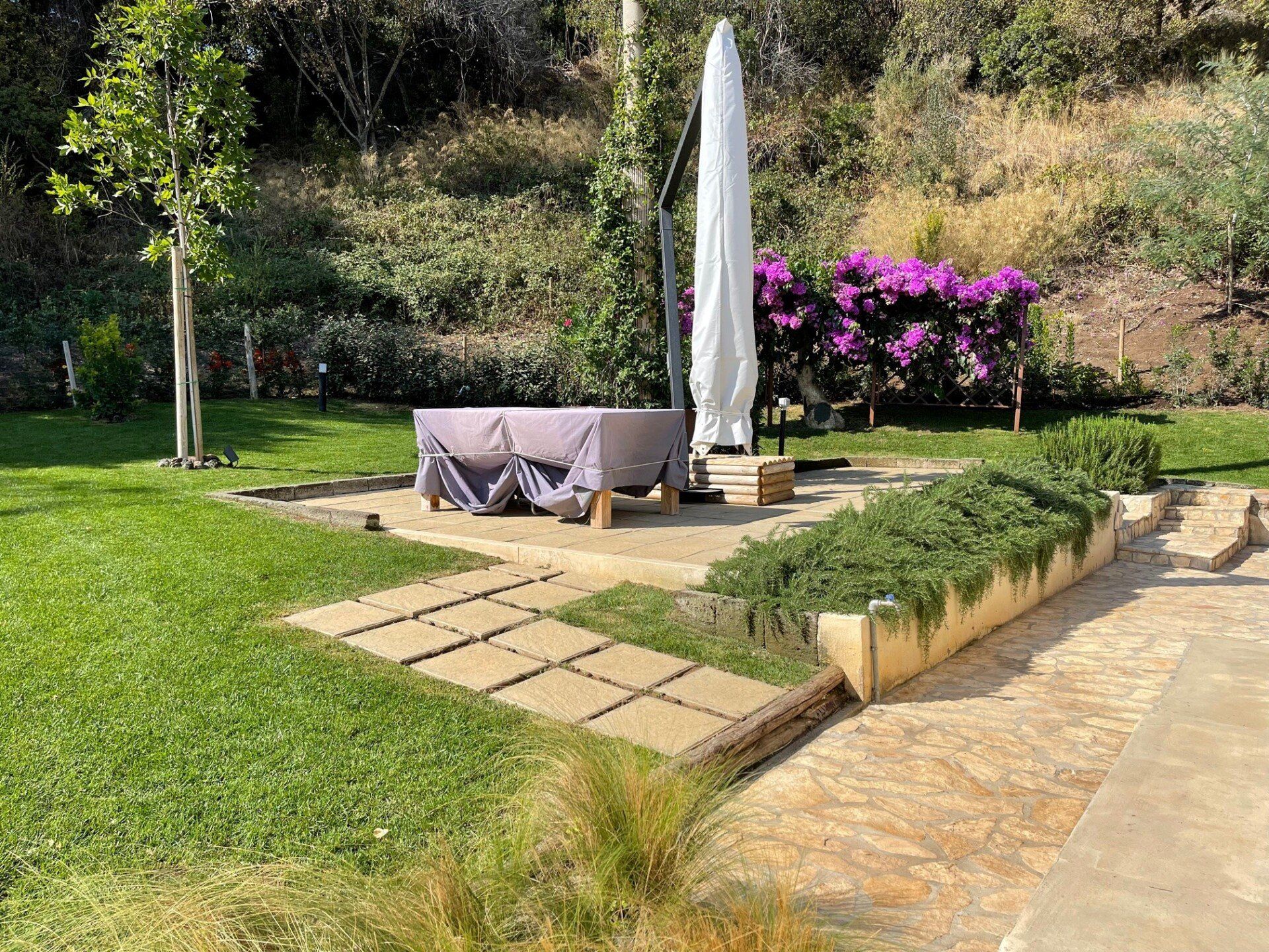 In un giardino soleggiato, un patio in pietra è arredato con un tavolo coperto, un ombrellone chiuso e una fioriera rialzata con arbusti verdi.