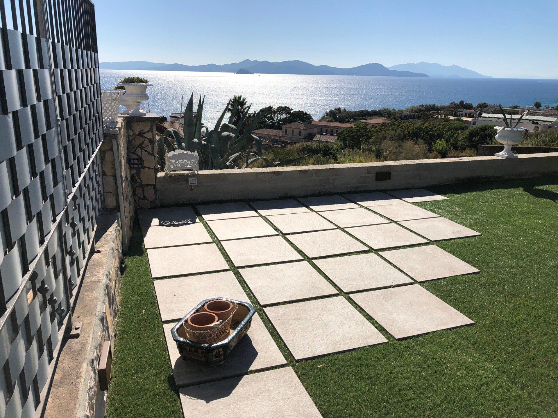 Un patio con pavimentazione di colore chiaro posata su erba artificiale, con vista sull'oceano e montagne sullo sfondo.