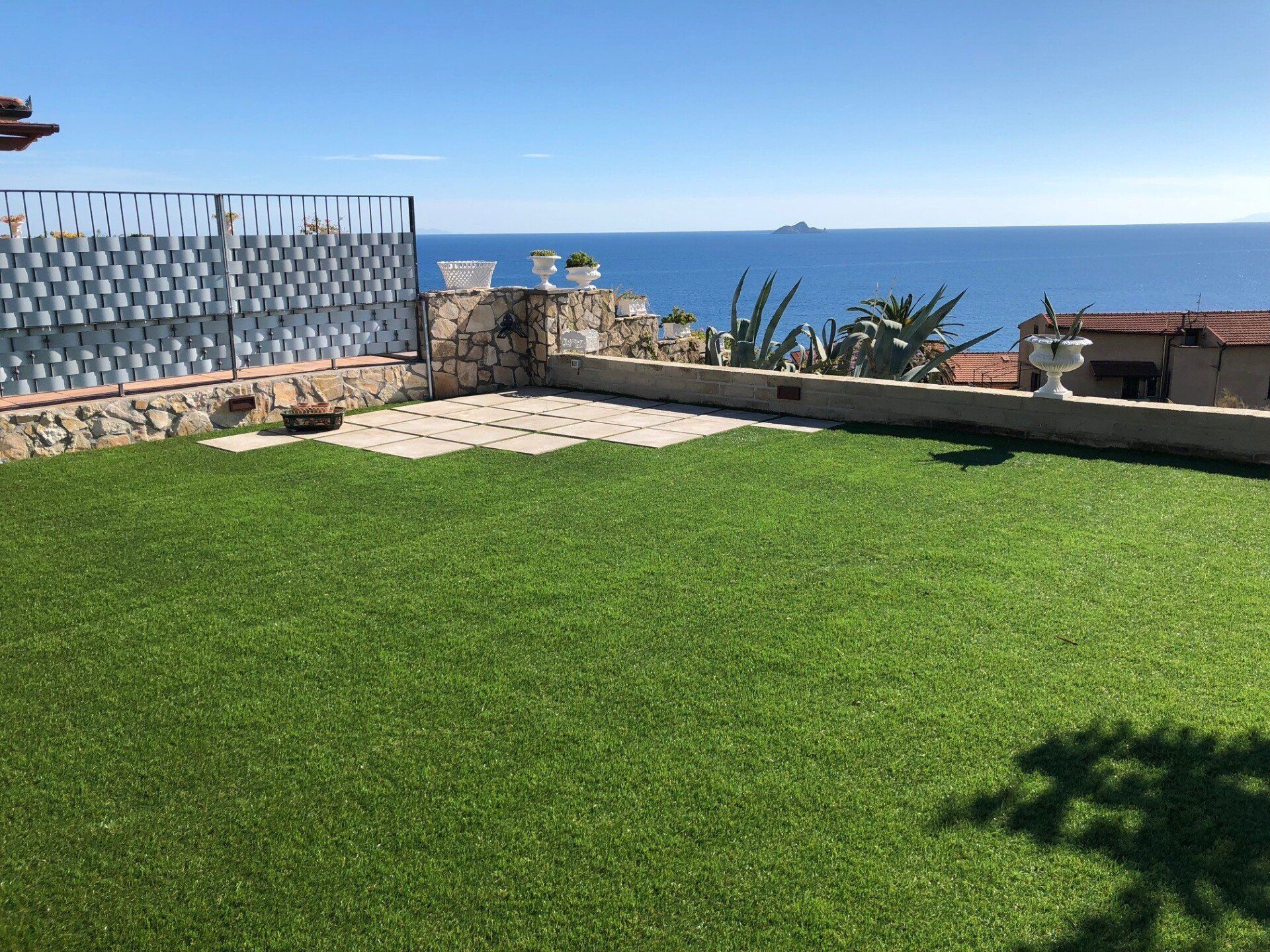 Una terrazza erbosa affacciata sul mare, delimitata da un muro di pietra, una recinzione metallica e la costa in lontananza, sotto un cielo limpido.