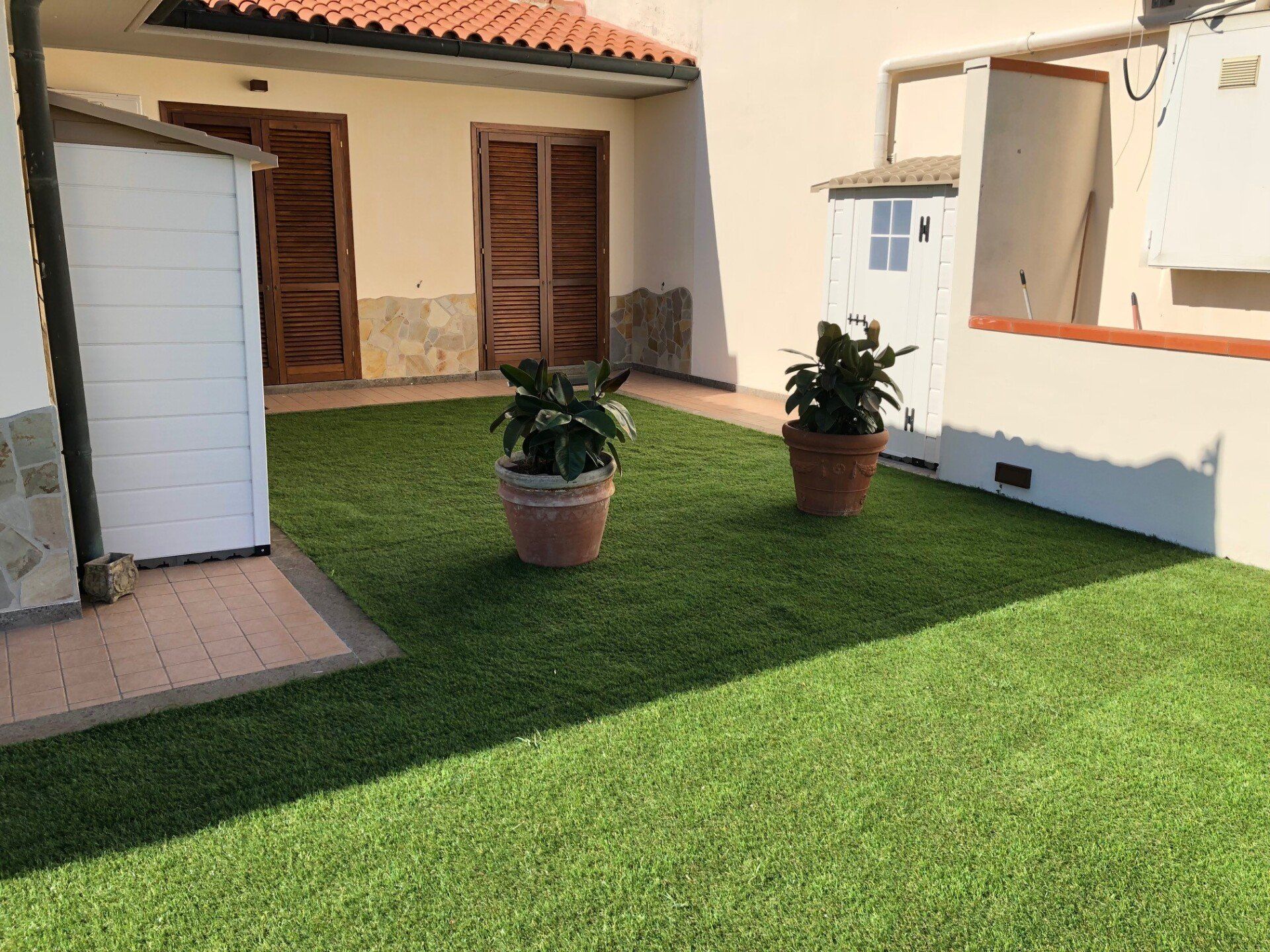 Un piccolo cortile erboso con due piante in vaso, un capanno bianco per gli attrezzi e un edificio con due porte di legno.