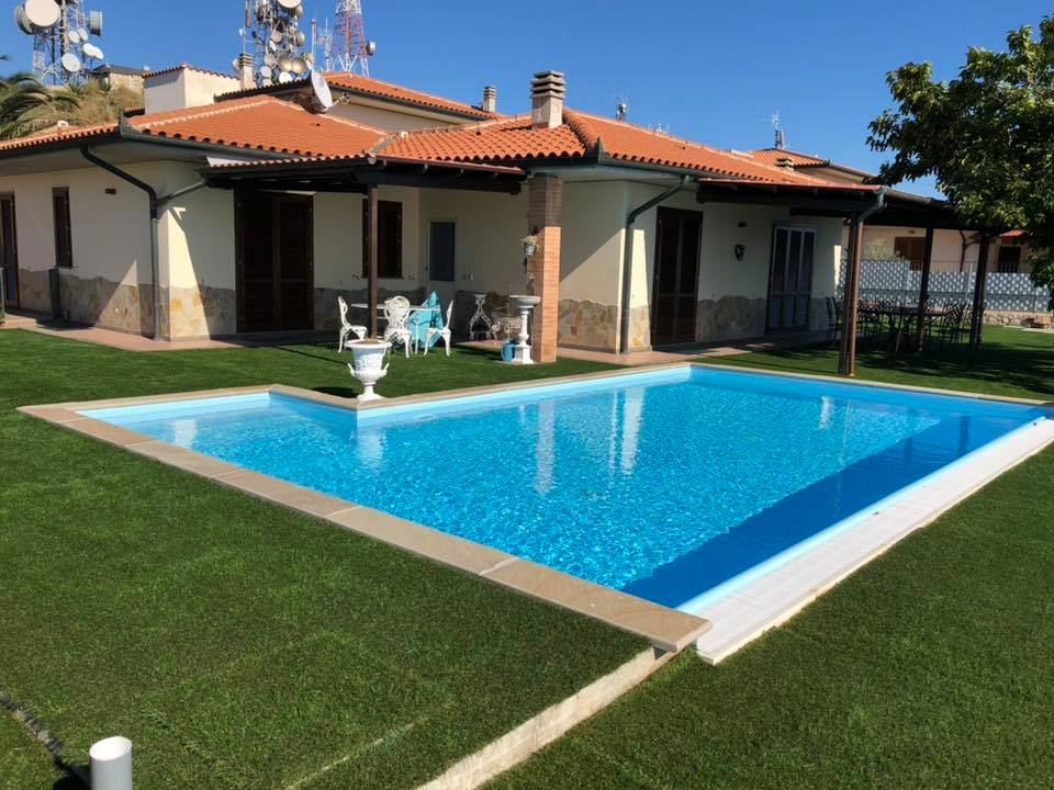 Una piscina a forma di L di un blu brillante si erge su un prato verde di fronte a una casa con tetto di tegole e patio coperto.