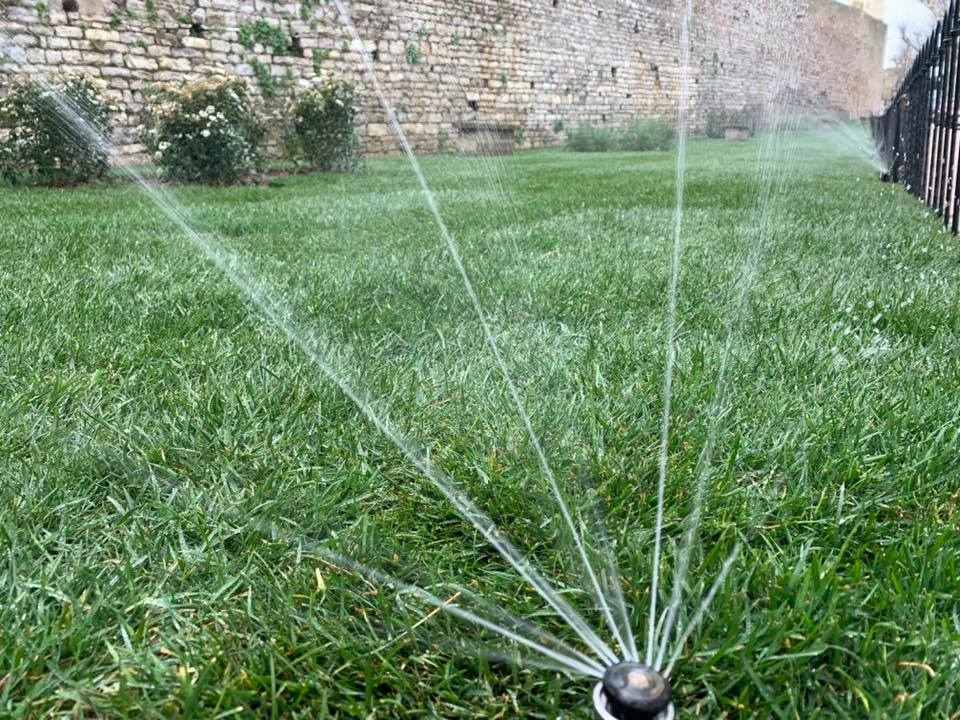 Un irrigatore da giardino spruzza getti d'acqua sull'erba verde di fronte a un muro di pietra.