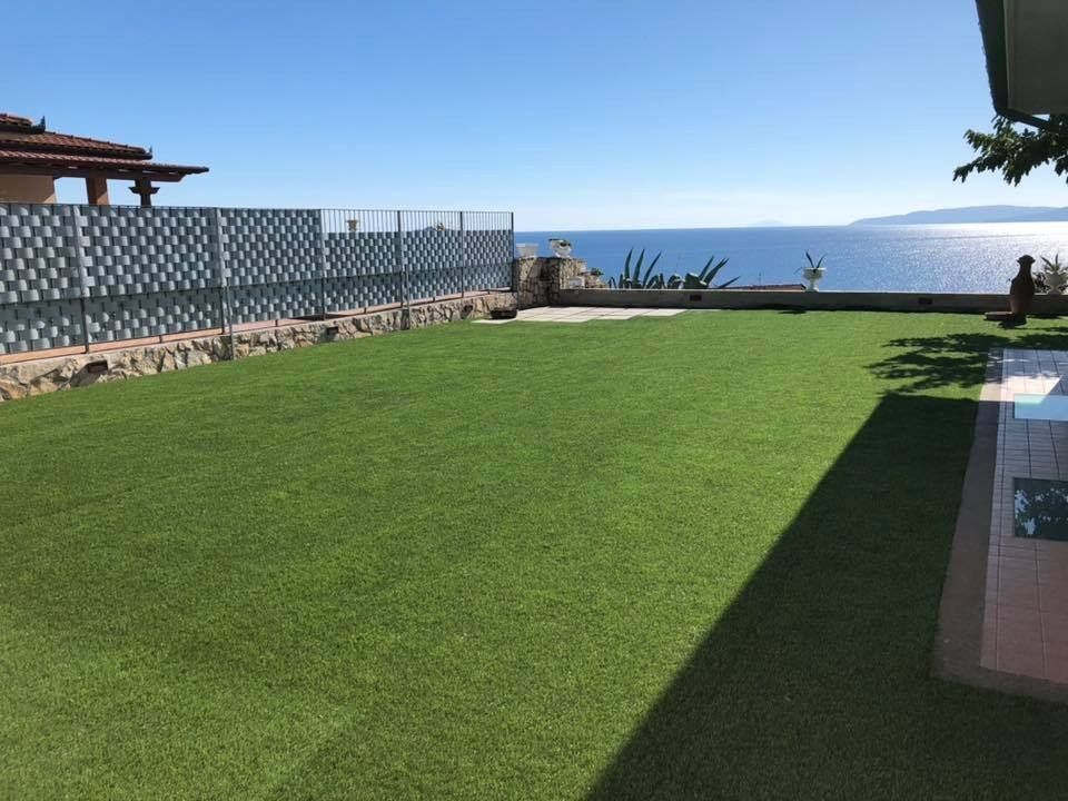 Un prato verde brillante che si affaccia sul mare blu sotto un cielo limpido, con una casa e un patio piastrellato in primo piano.