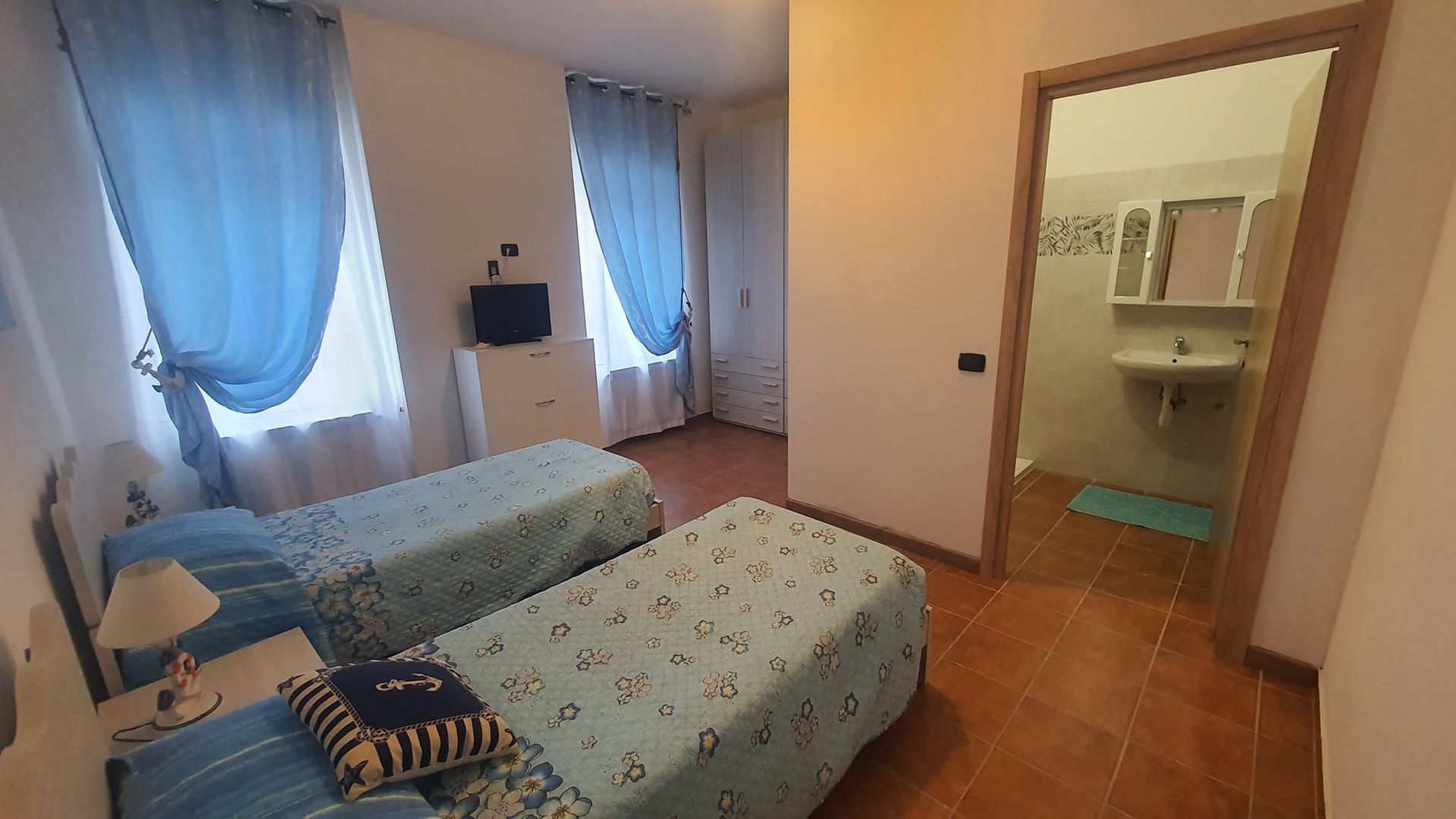 Due letti singoli in una camera da letto con tende blu, cassettiera e una porta aperta sul bagno.
