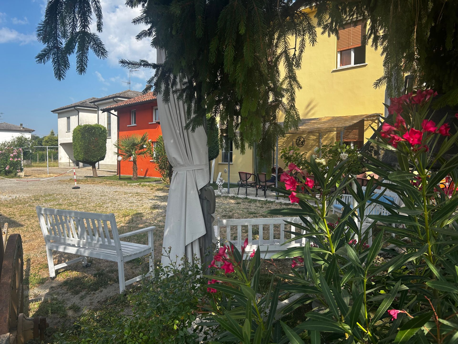 Giardino con panchine bianche, fiori ed edifici colorati sullo sfondo sotto un cielo azzurro.