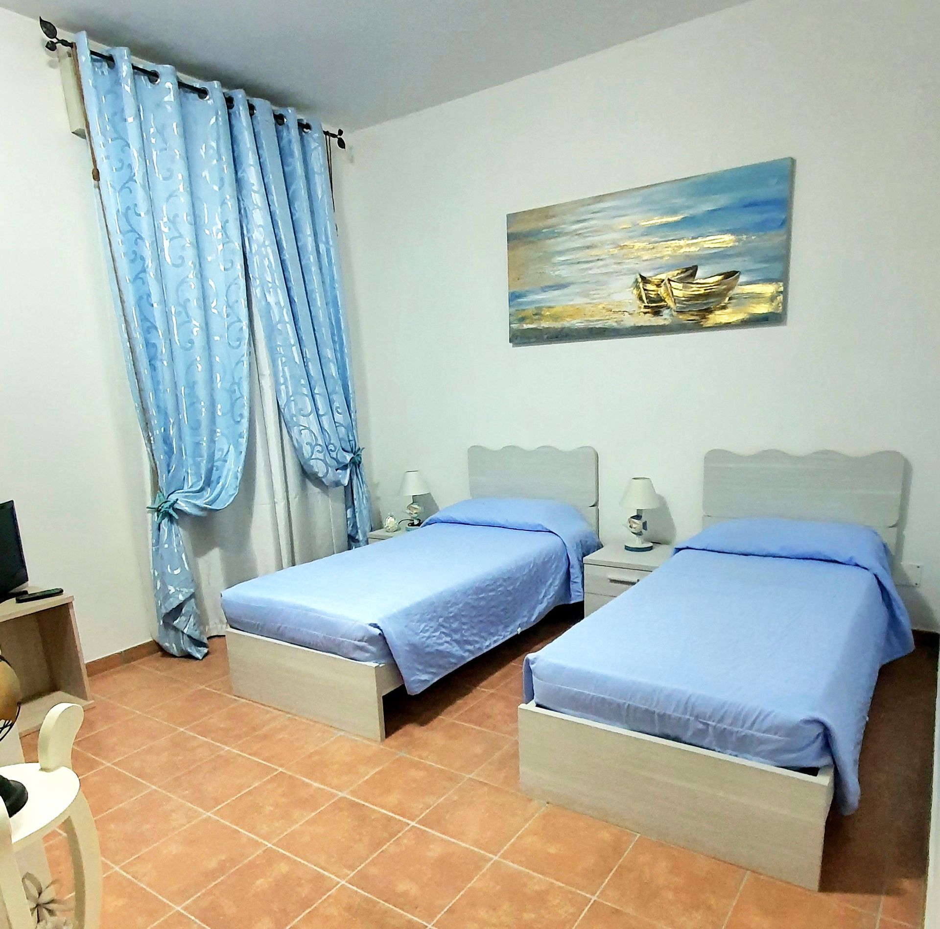Camera da letto con due letti singoli, biancheria da letto azzurra e un dipinto di barche; tende blu.