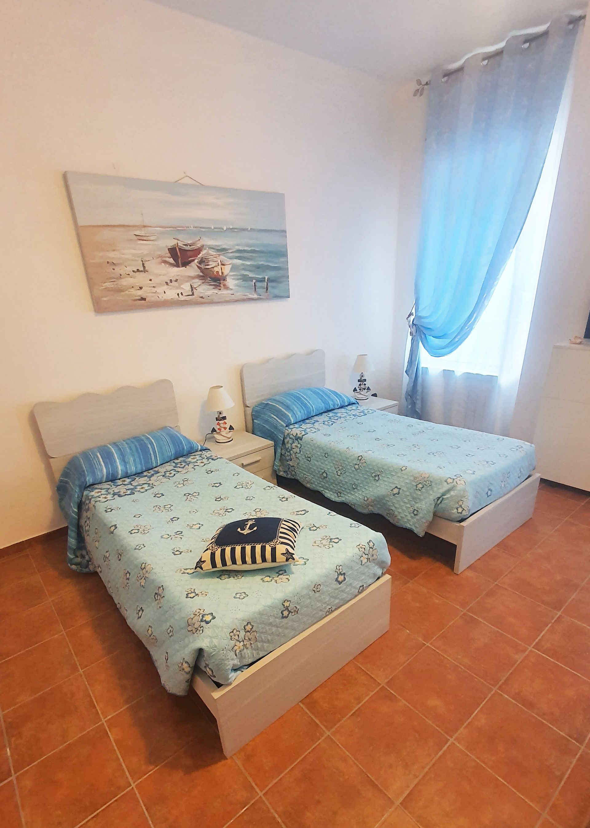 Due letti singoli in una stanza con un dipinto, tende blu e pavimento piastrellato arancione.