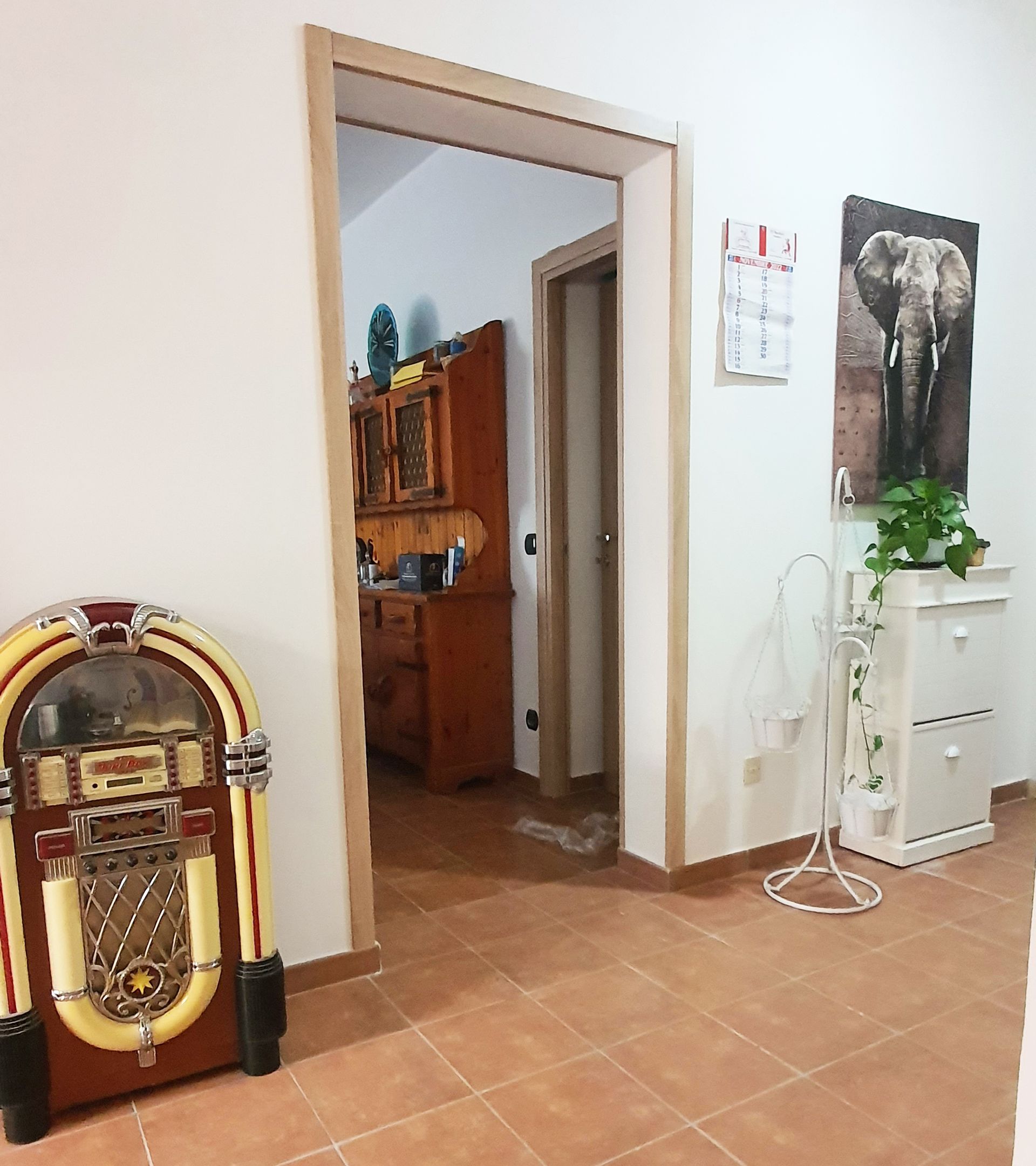 Jukebox vicino a una porta che conduce a un mobiletto di legno. Vicino alla porta si trovano un dipinto di un elefante e un mobiletto bianco.