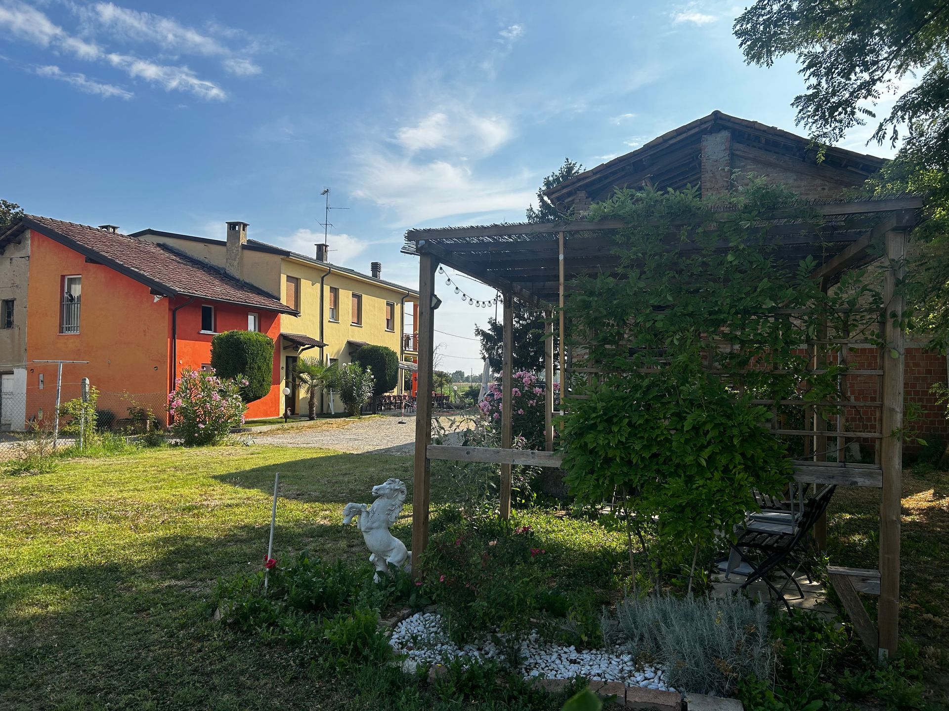 Un cortile con un pergolato in legno ricoperto di viti, di fronte a case colorate sotto un cielo azzurro.