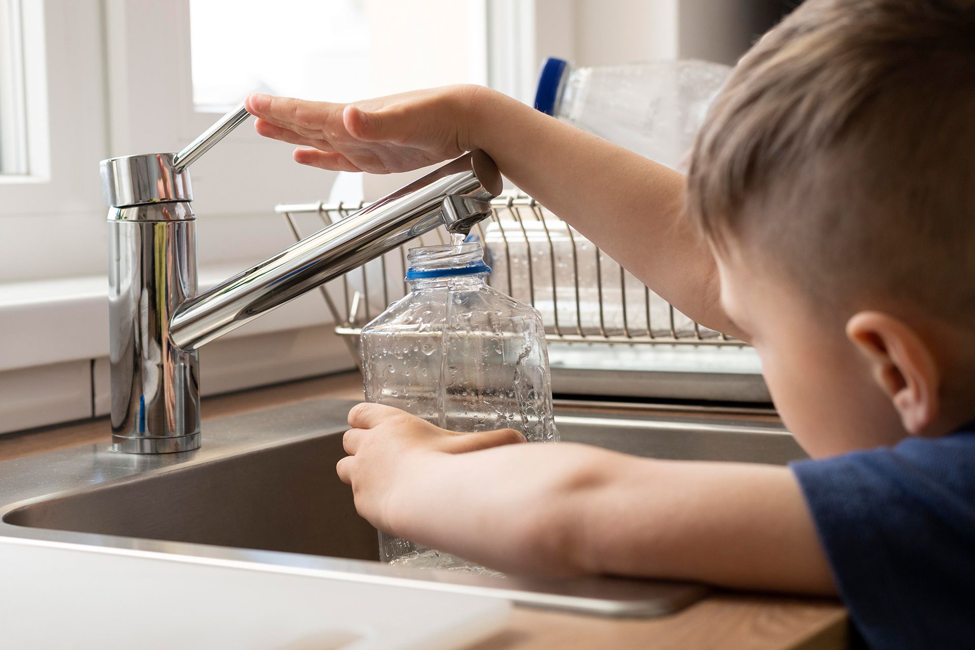 Child filling a clear plastic water bottle from a chrome faucet in a kitchen sink.