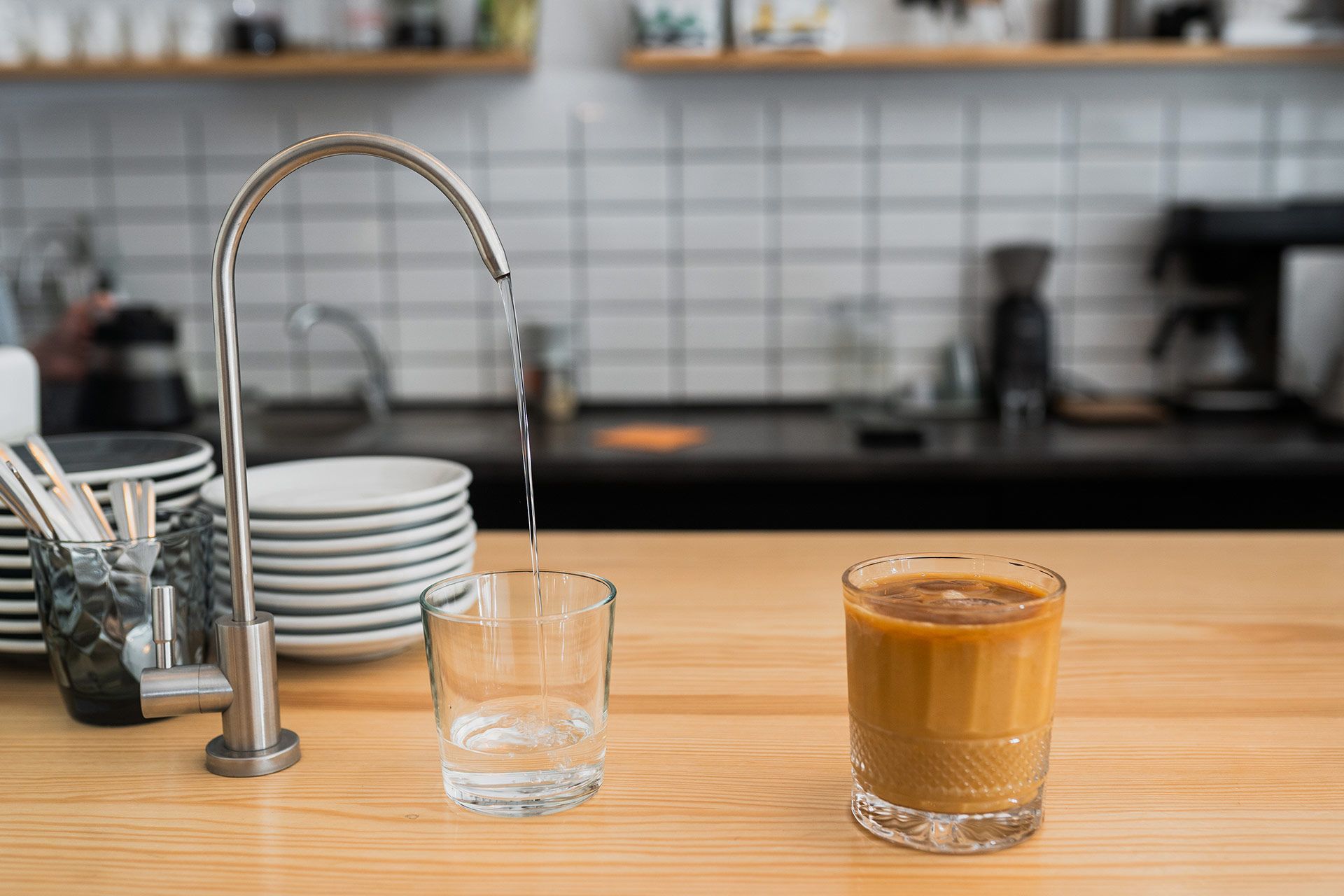 Water pouring into a clear glass, next to a glass of coffee on a wooden counter in a cafe.