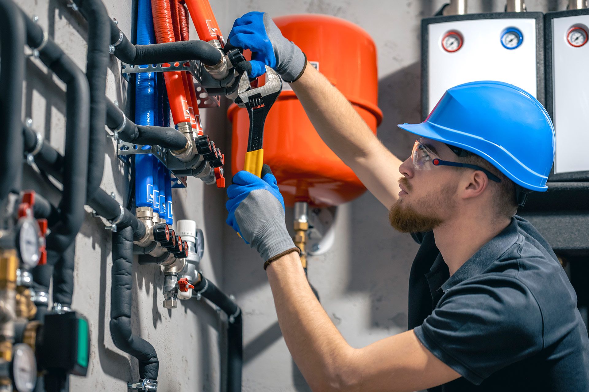 Plumber in a blue hard hat using pliers on pipes, in a utility room.