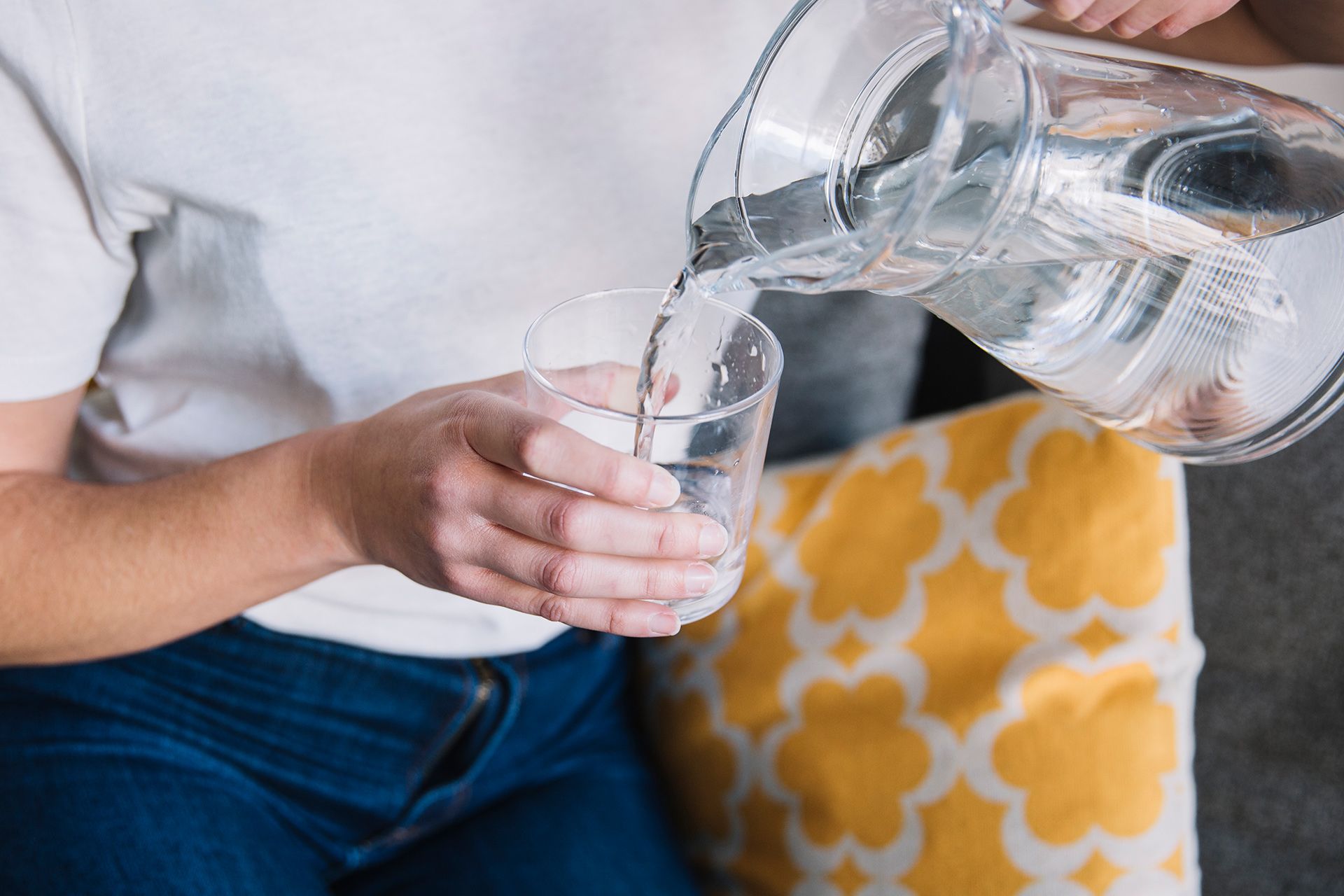 Person pouring water from a pitcher into a glass, sitting near a yellow patterned pillow.