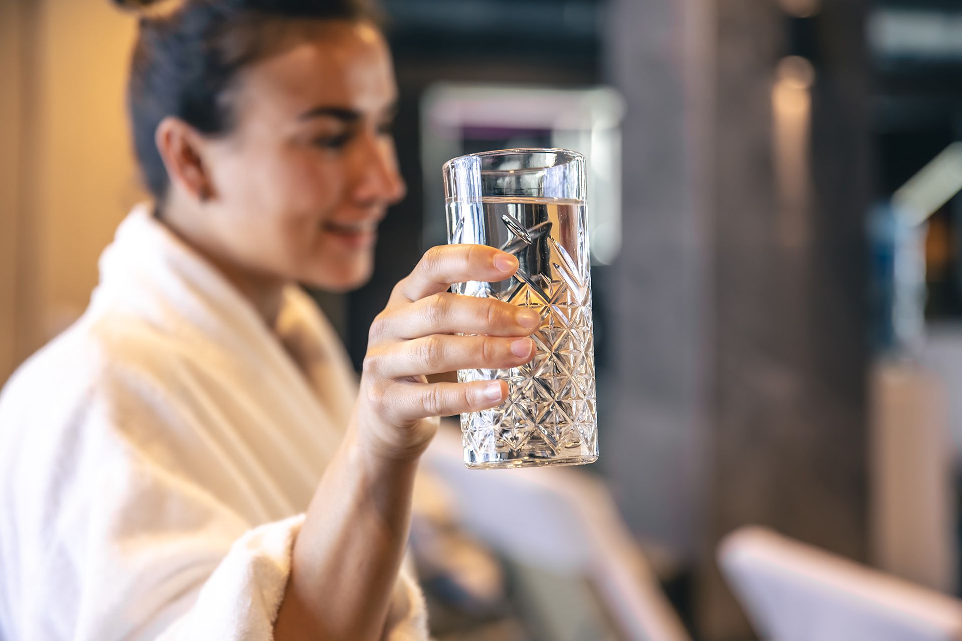 Woman in white robe smiling, holding a decorative glass of water in a spa setting.
