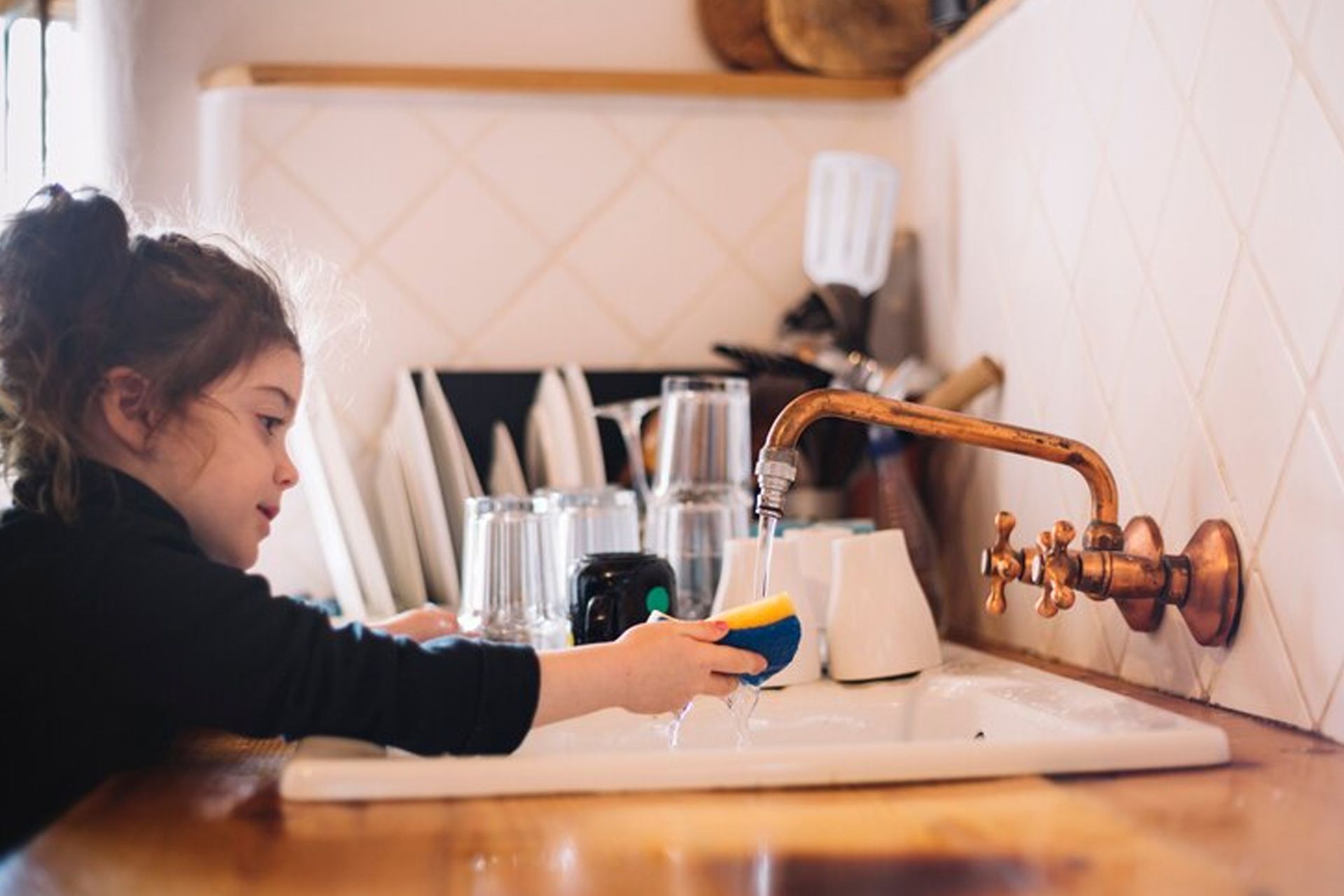 Young child washing a sponge in a kitchen sink. Water runs from copper faucet.