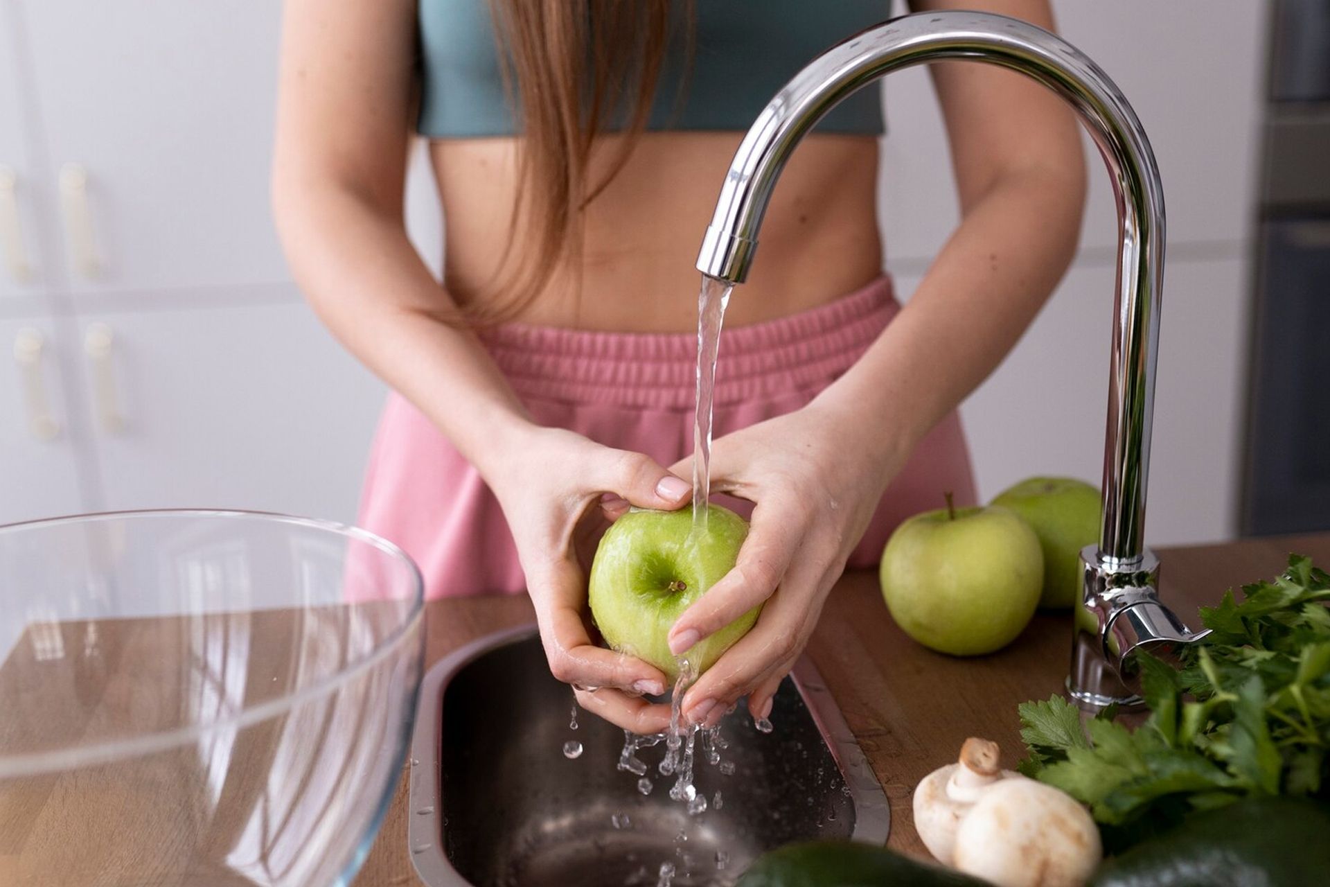 Woman washes a green apple under a faucet in a kitchen sink.