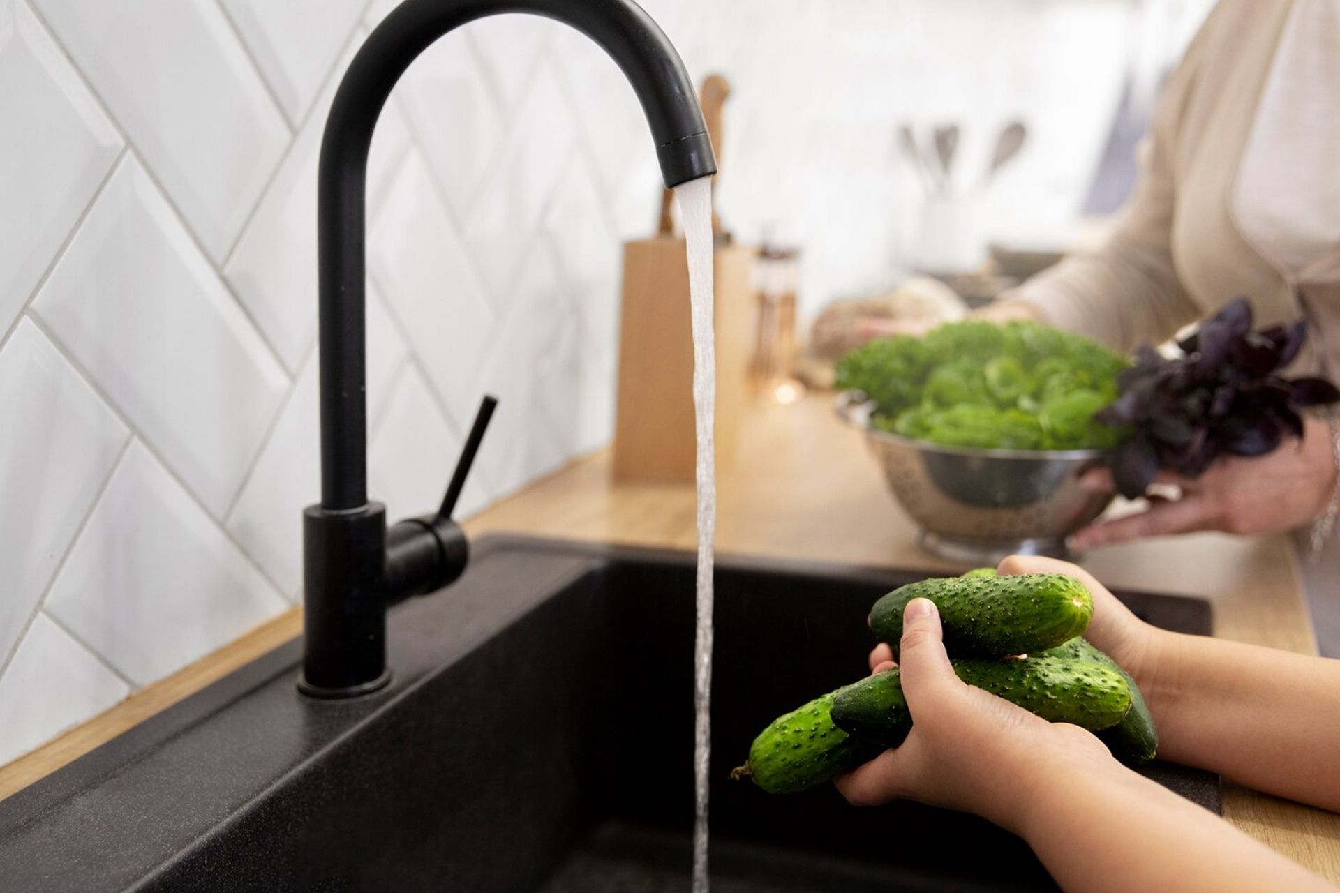 A person washing cucumbers in a black sink under a running faucet. Other vegetables are nearby.
