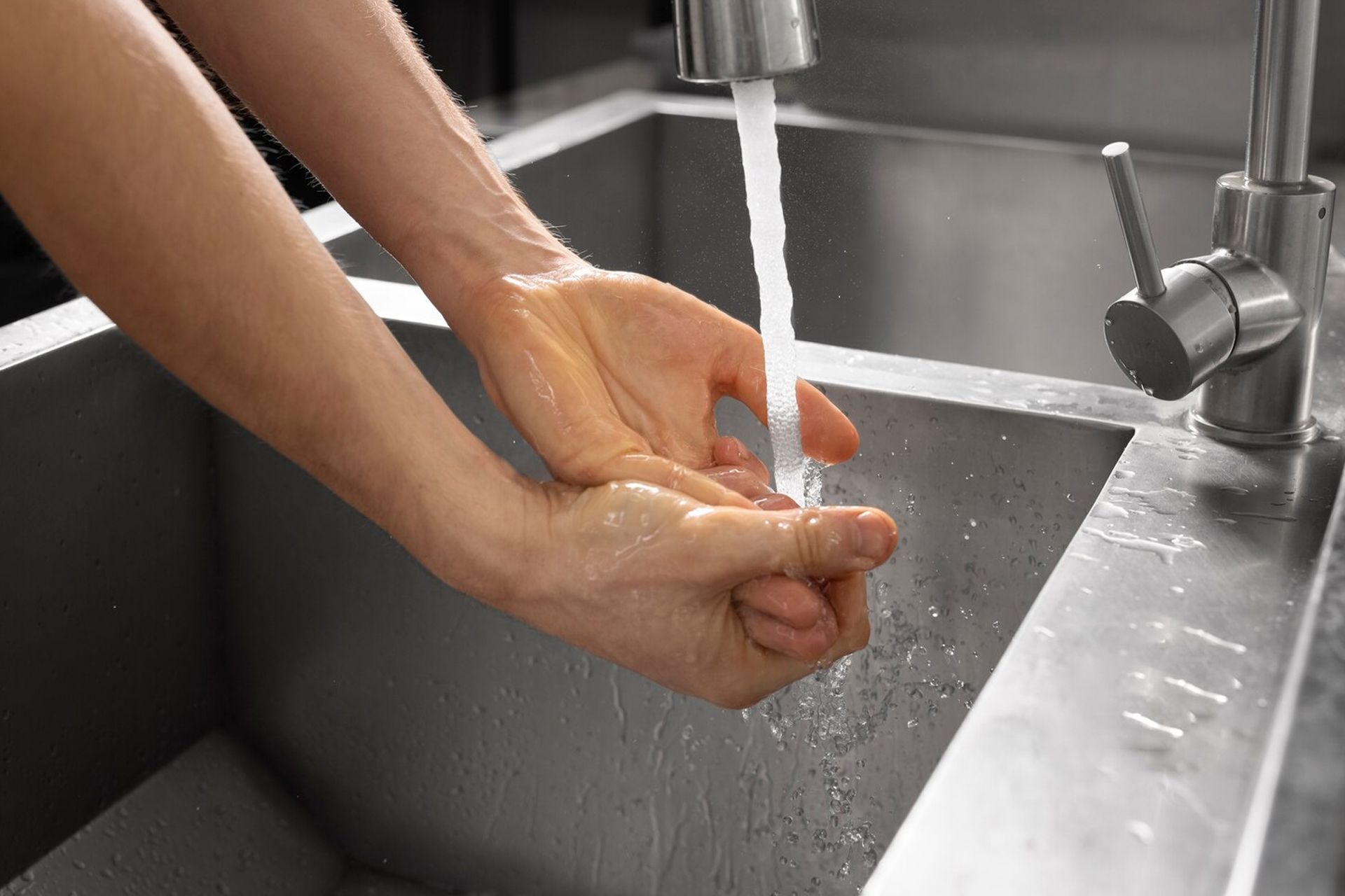 Hands under running water in a stainless steel sink, person washing hands.