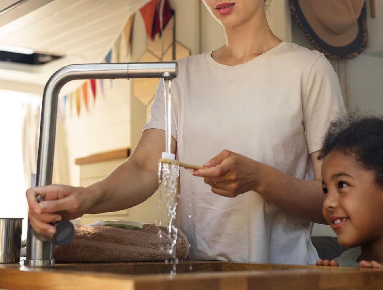 Woman rinsing a toothbrush under a faucet, child watches, bright kitchen.