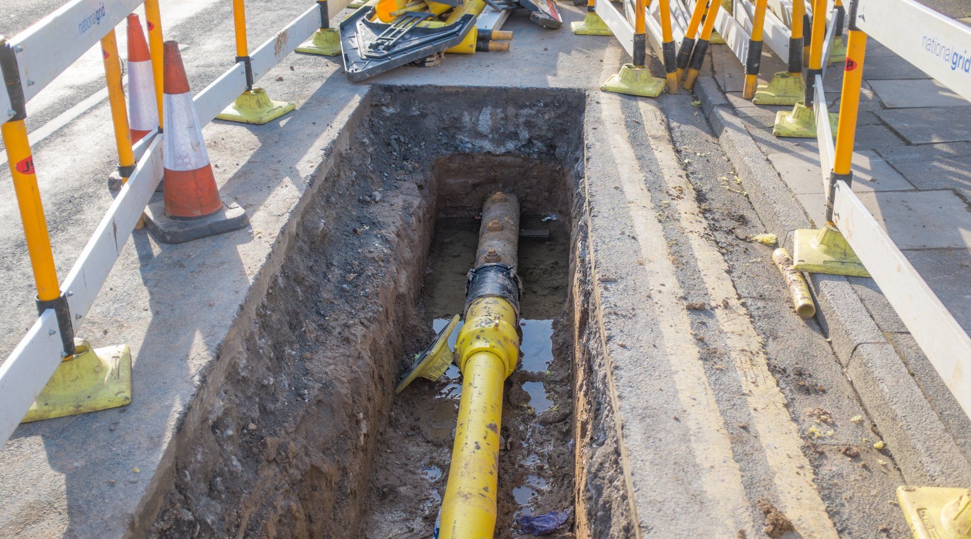 A trench in a city street reveals a yellow pipe connection surrounded by construction barriers and orange safety cones.