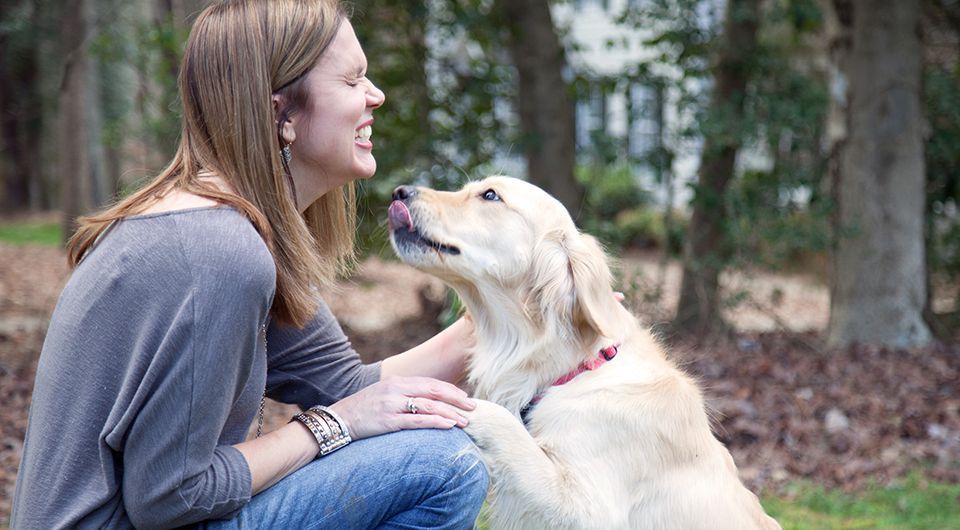 Woman kneels outdoors, smiling at a cream-colored dog wearing a pink collar and nuzzling her hand.