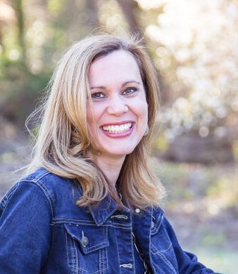 Smiling blonde woman in a denim jacket outdoors with a blurred natural background