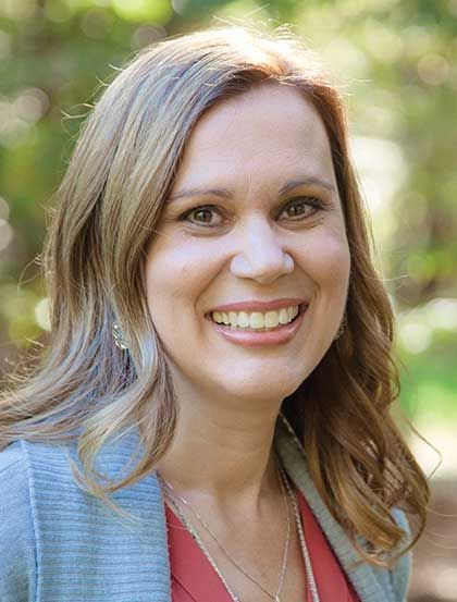 Smiling woman outdoors with blonde hair, wearing a blue cardigan and coral top, against a blurred green background