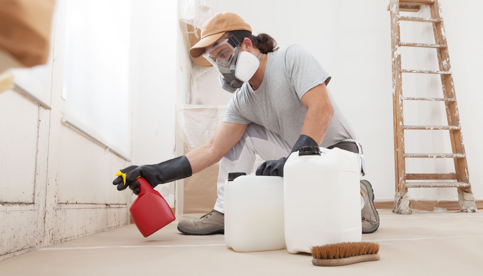 Worker in mask and goggles spraying mold treatment on floor during home remediation