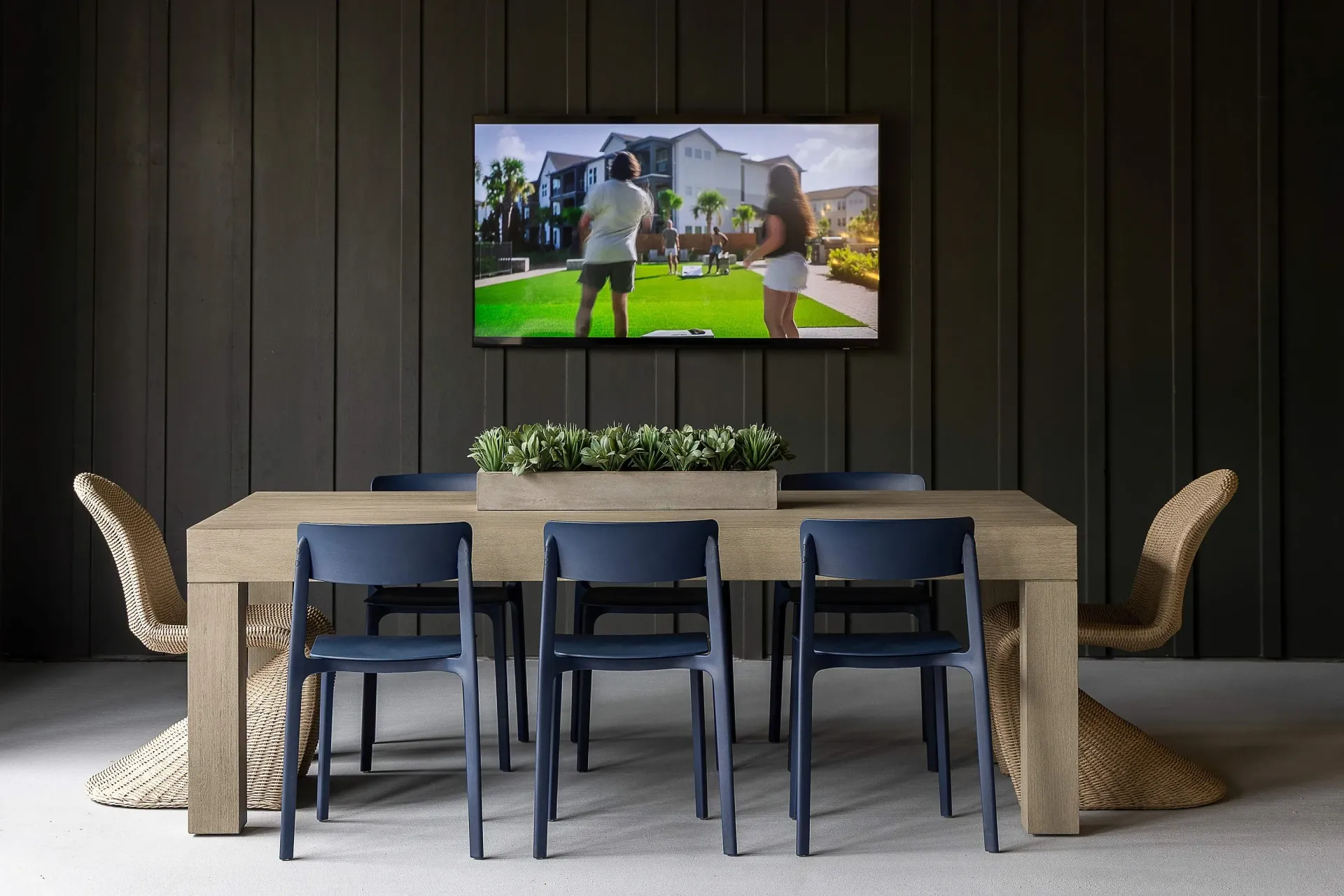 A large table with chairs is set up in front of a TV displaying an outdoor scene with people playing lawn games.