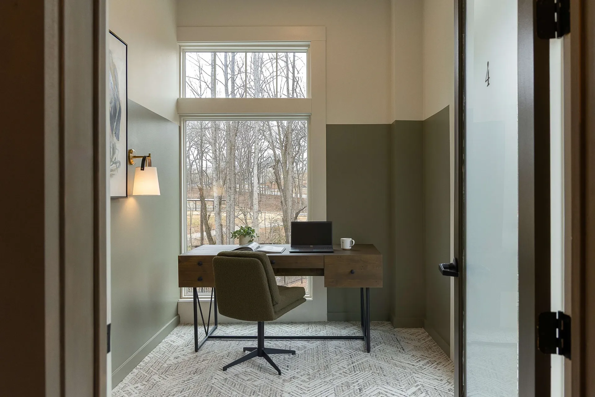 A home office with a desk, chair, laptop, and mug overlooking trees.