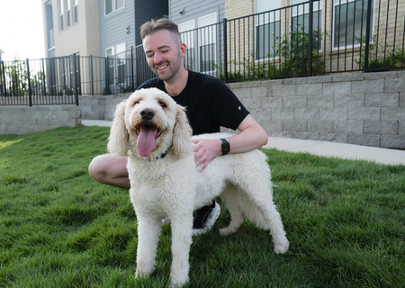 Man crouches beside a fluffy white dog on green grass outside an apartment building.