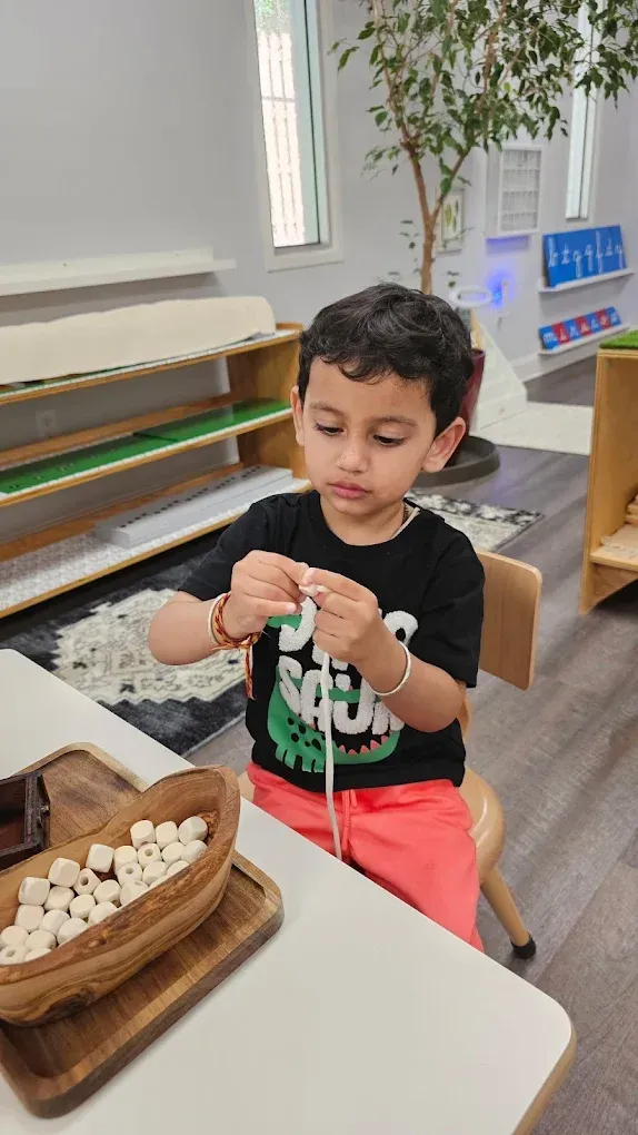 Child stringing beads at a table in a classroom, with wooden trays and a plant in the background