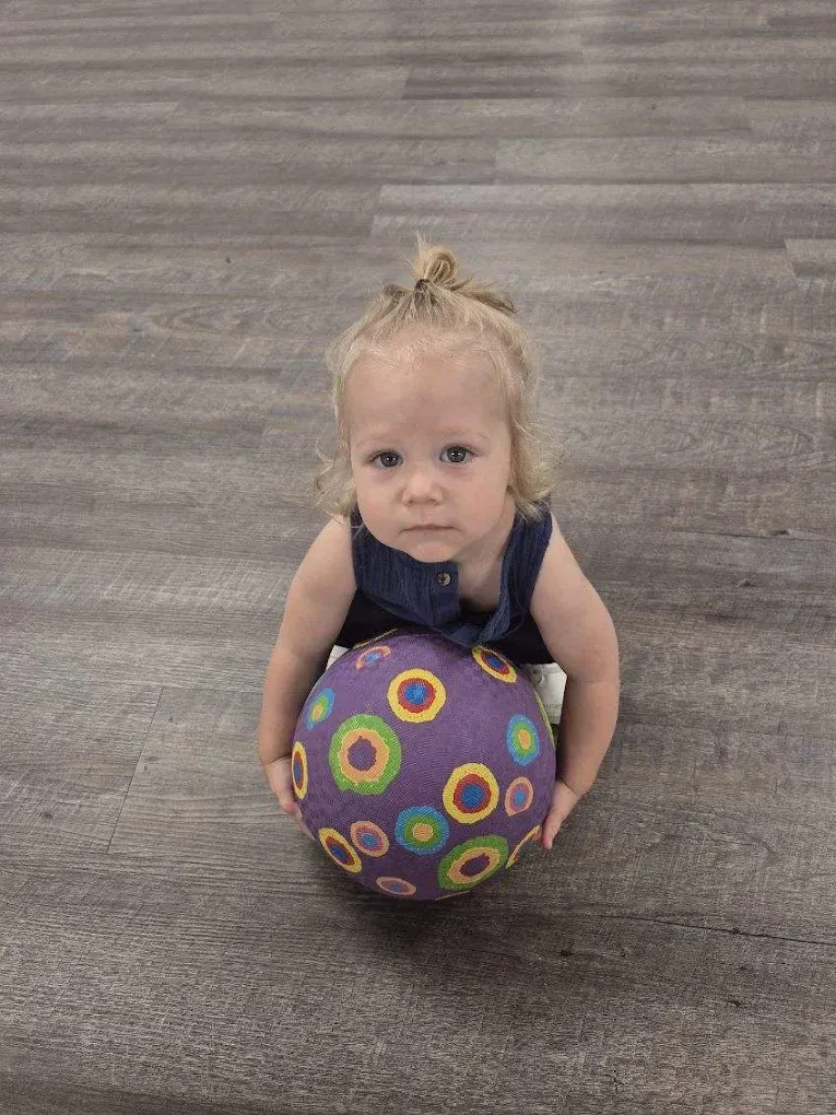 Toddler sitting on a gray floor, holding a colorful patterned ball and looking at the camera.