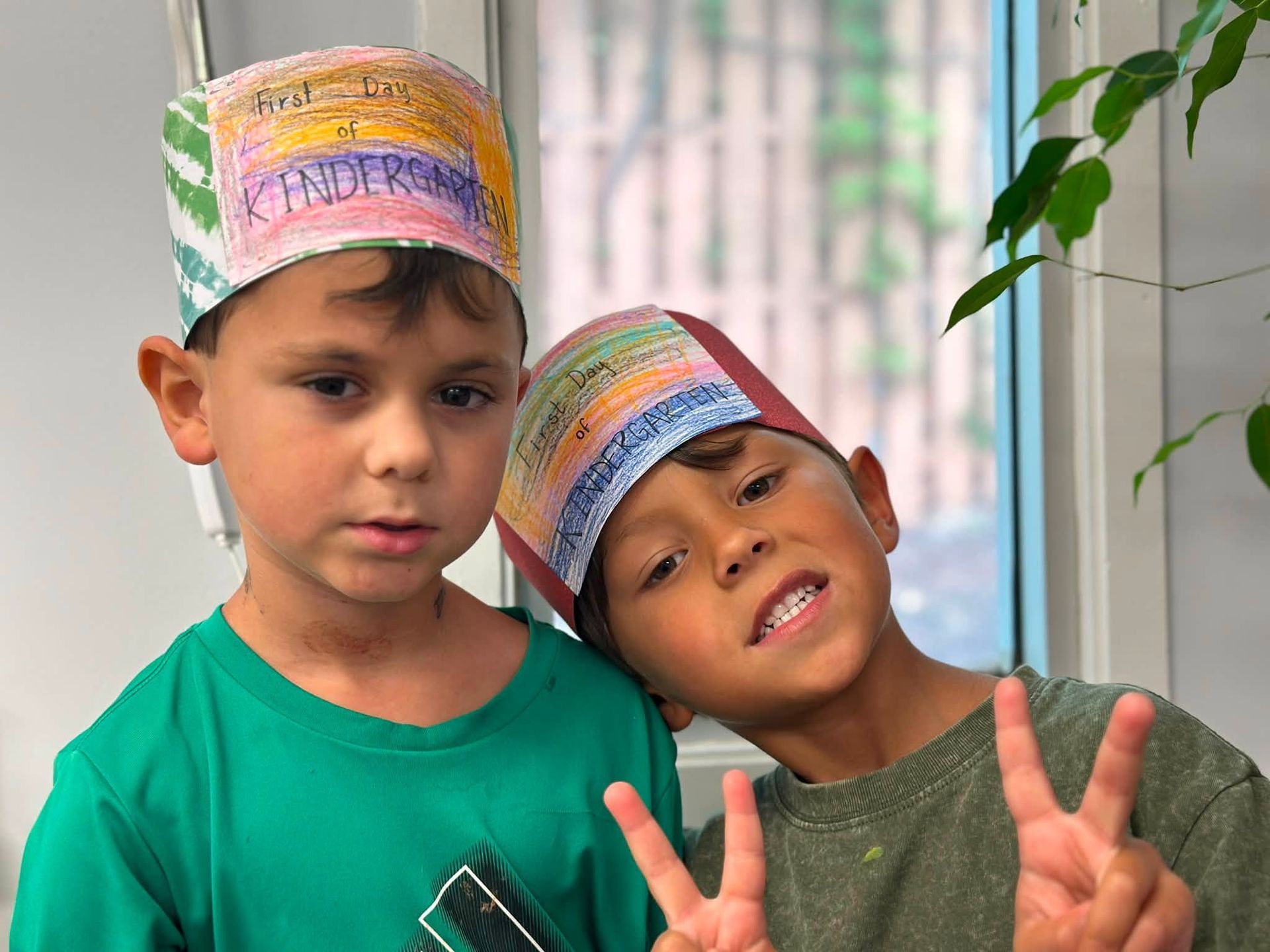 Two children wearing colorful paper hats, one smiling and flashing peace signs indoors.