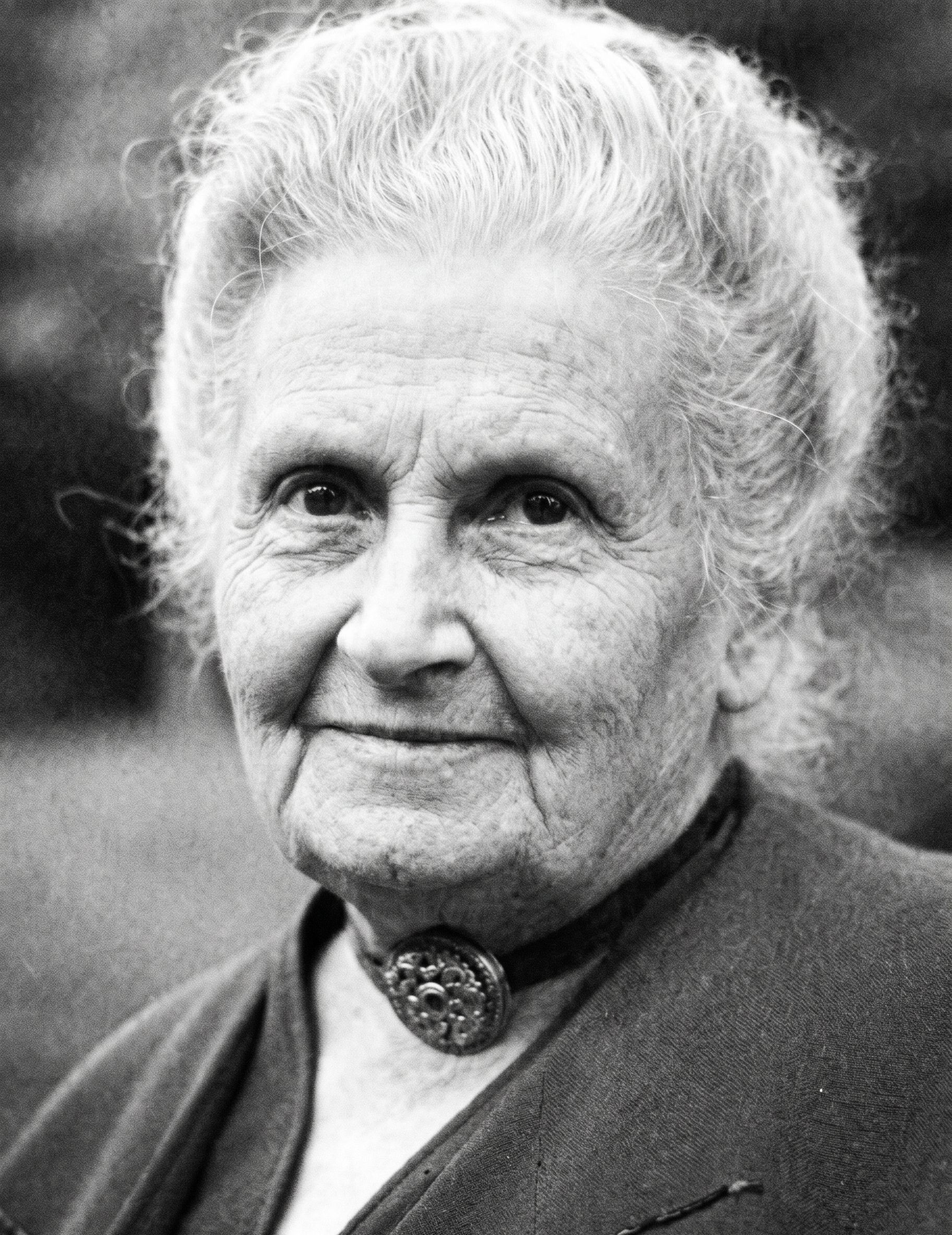 Black-and-white portrait of an elderly woman with a gentle smile, wearing a dark jacket and brooch.
